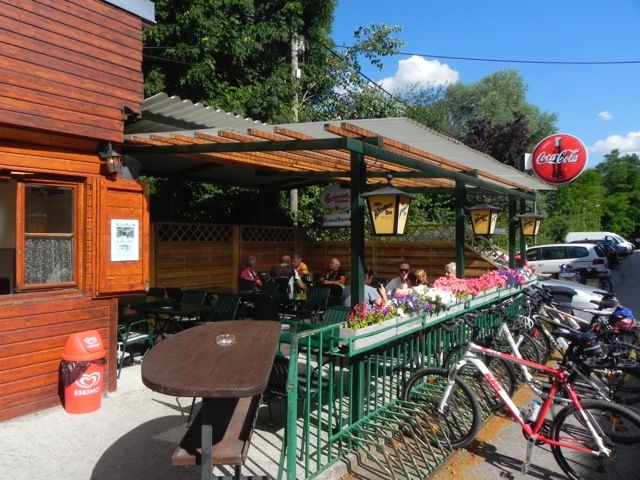 Außenbereich eines Imbisses mit Holzwänden, Tischen und Fahrrädern. Menschen sitzen unter einem überdachten Bereich. Coca-Cola-Schild sichtbar.