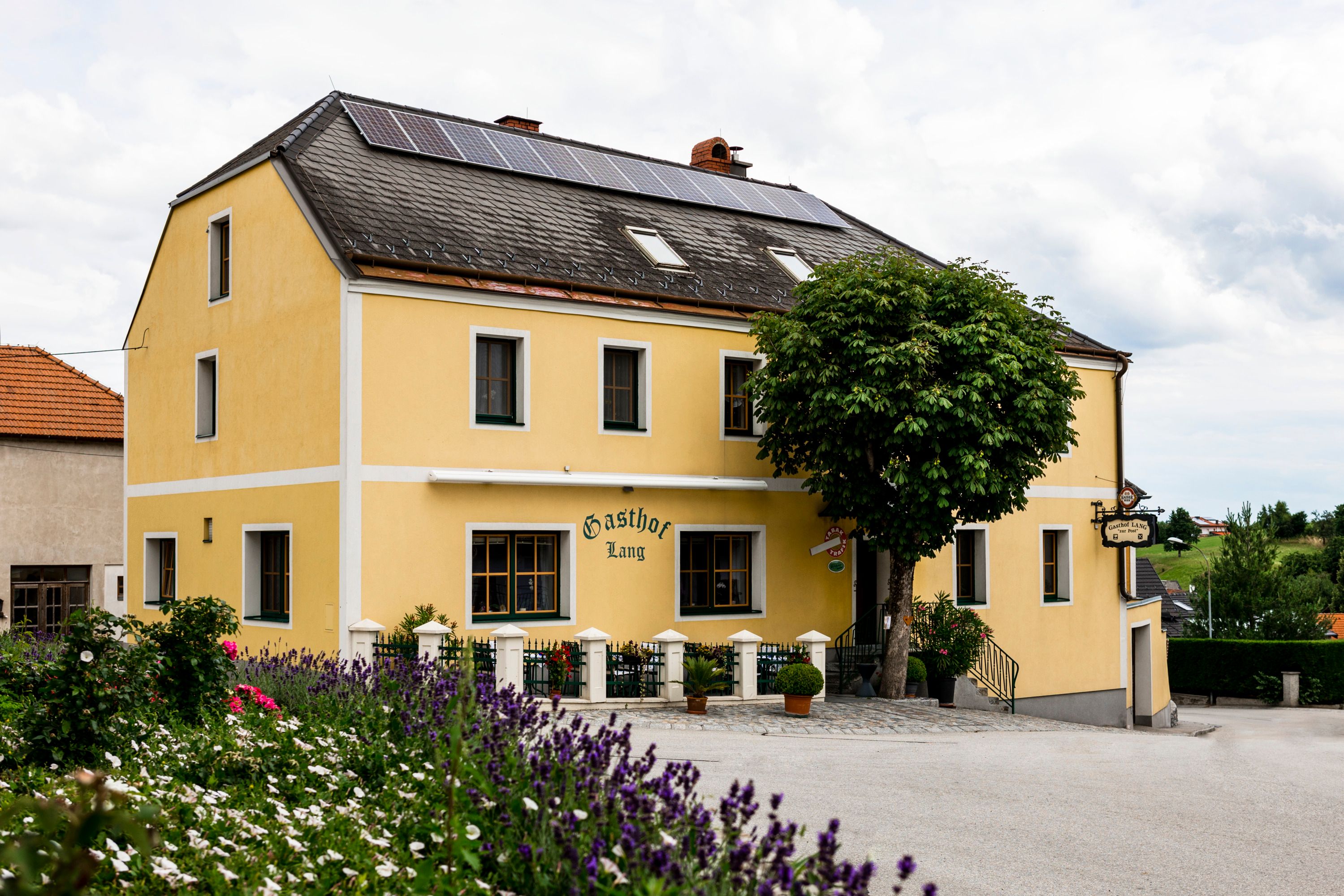 Gelbes Gasthaus mit Solarpanelen auf dem Dach, umgeben von Blumen und Bäumen.