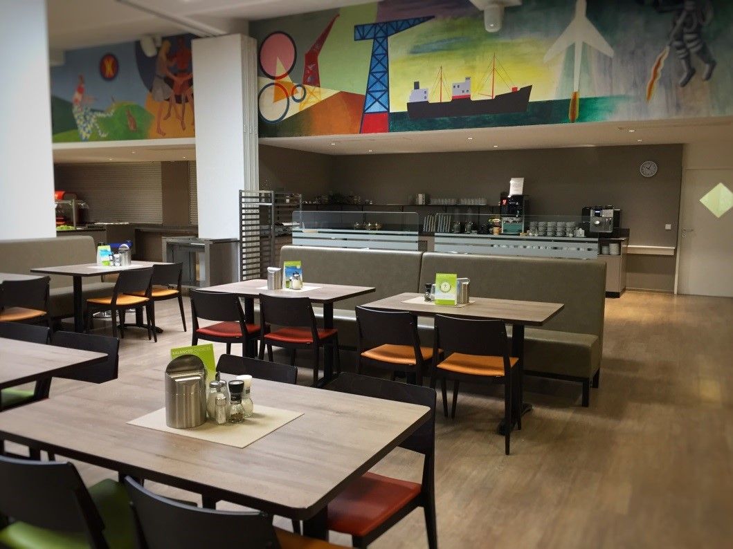 Innenansicht eines Restaurants mit leeren Tischen und Stühlen, bunte Wandmalerei im Hintergrund.