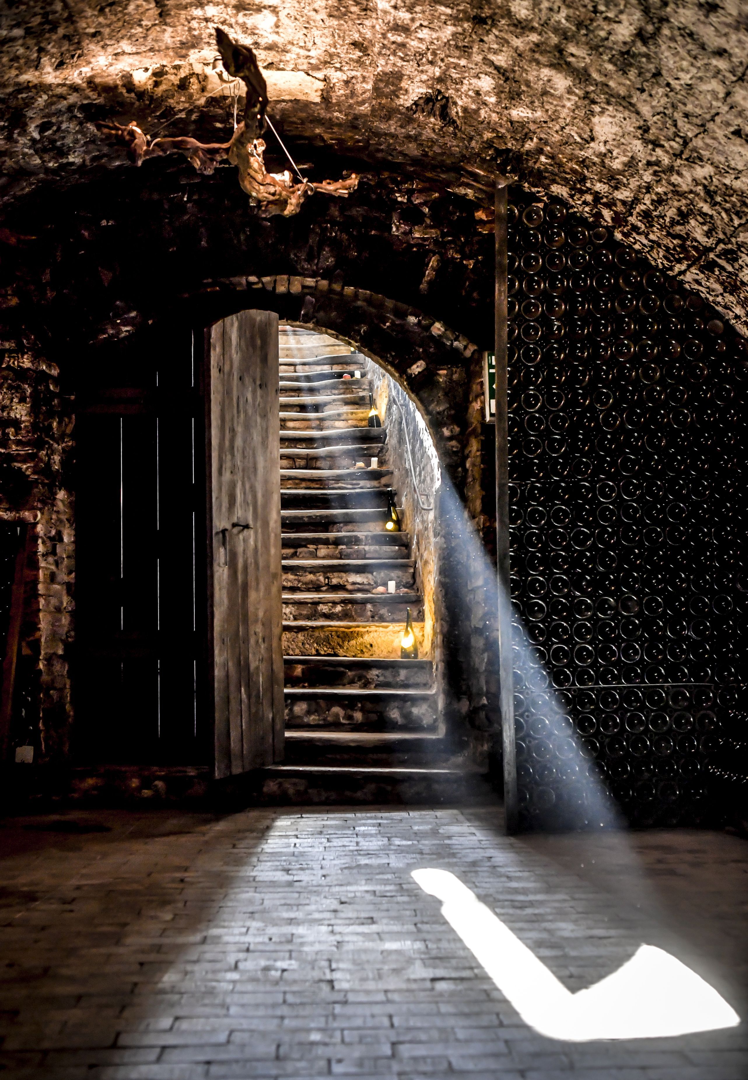Steinerne Treppe in einen Weinkeller mit Gewölbedecke, Flaschenwand und Lichtstrahl im Raum.