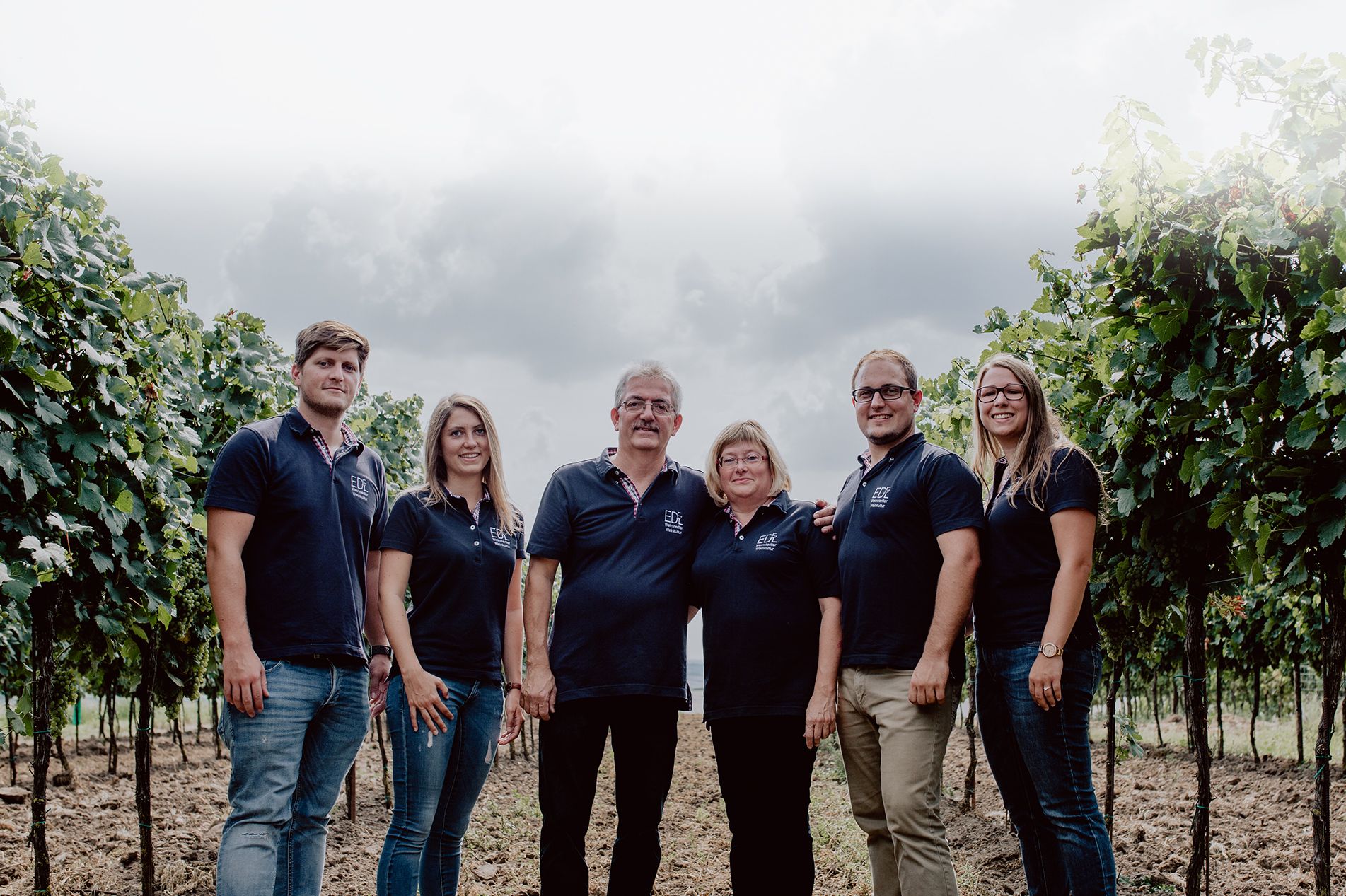 Eine Familie steht in einem Weinberg, alle tragen dunkelblaue Poloshirts mit einem Logo.
