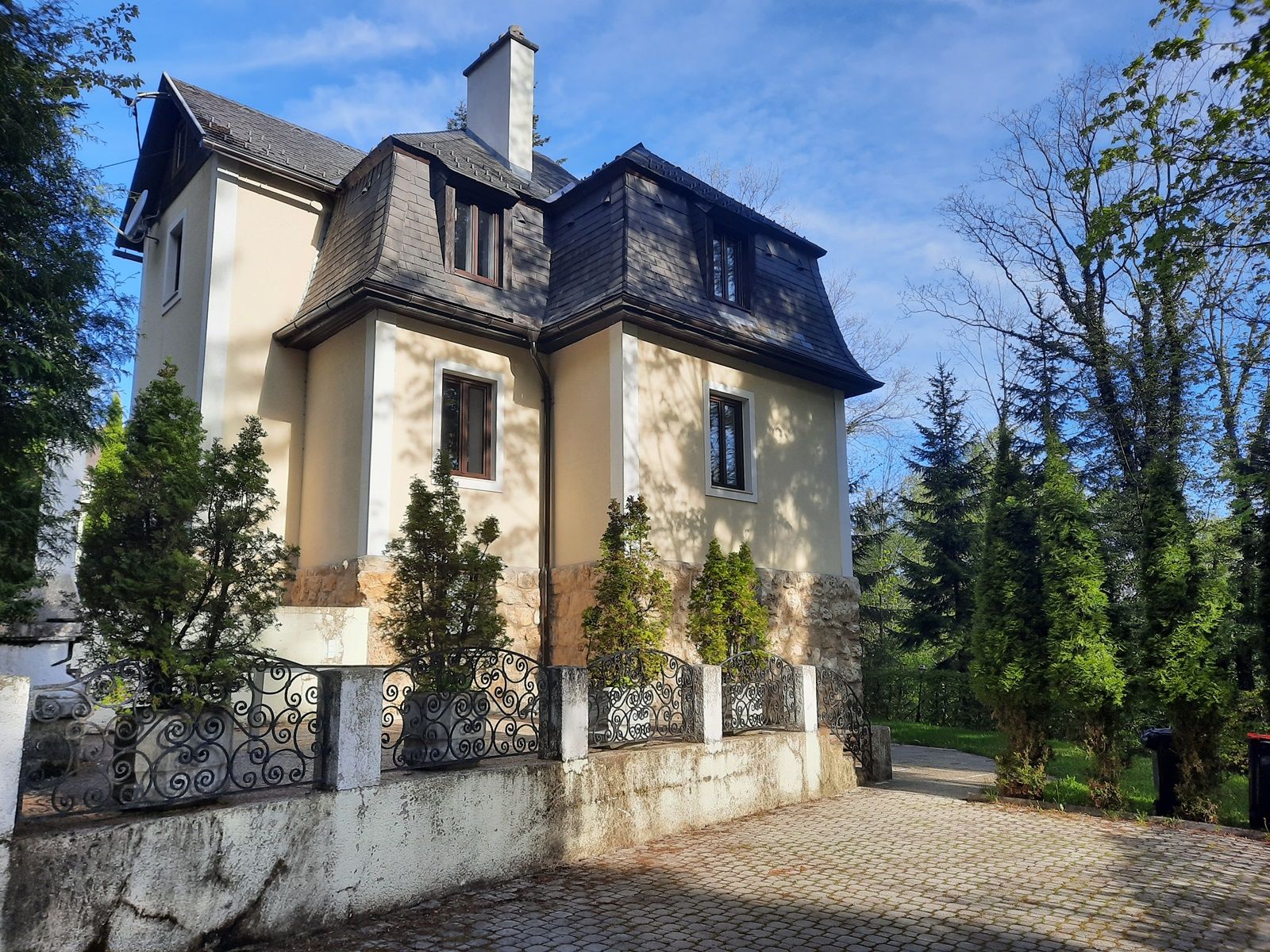 Eine zweistöckige Villa mit Mansardendach, umgeben von Bäumen und einem schmiedeeisernen Zaun.