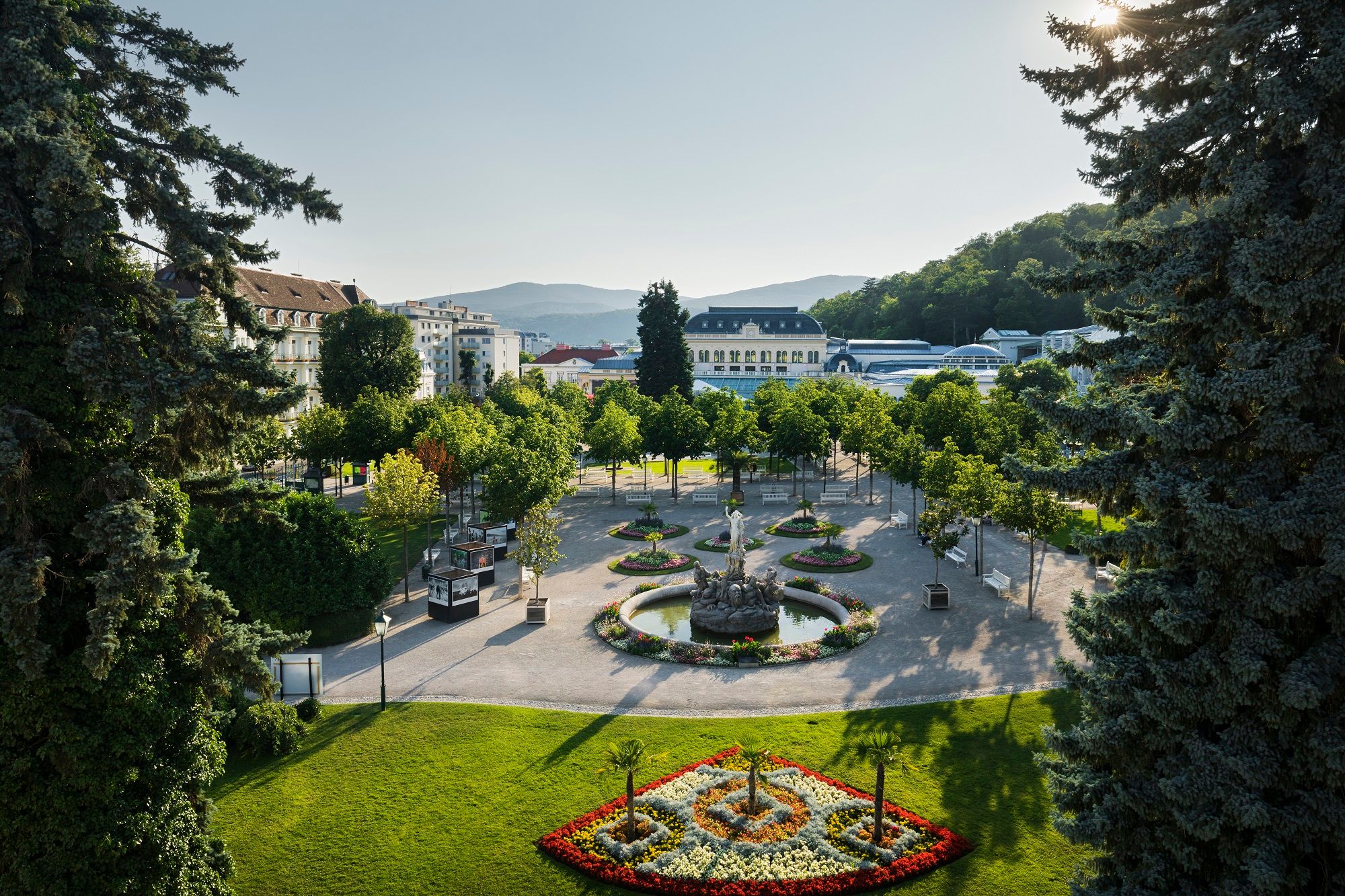 Ein gepflegter Park mit Blumenbeeten, Bäumen und einem Brunnen, umgeben von Gebäuden und Hügeln im Hintergrund.