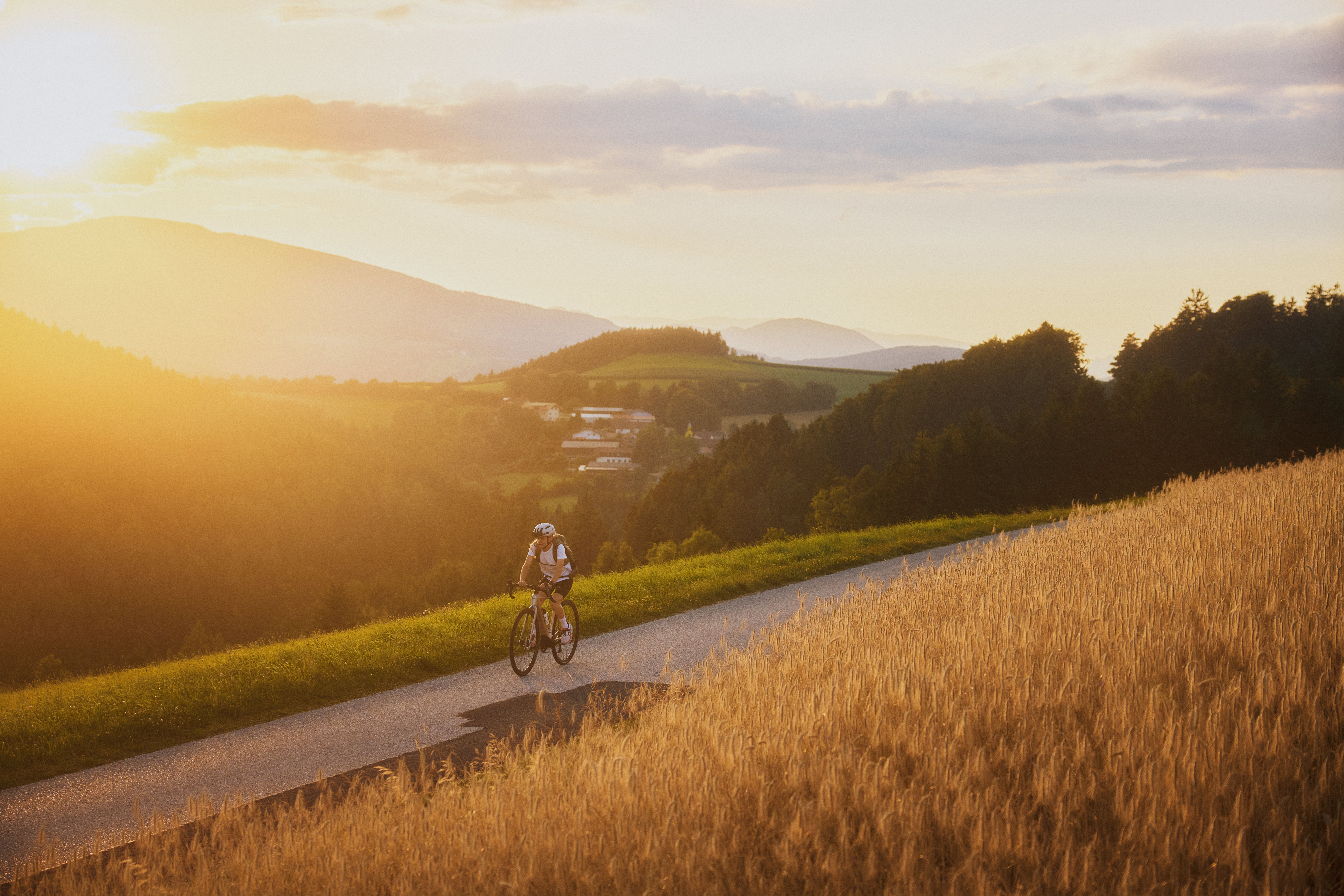 Eine Radfahrerin fährt bei Sonnenuntergang auf einer Landstraße durch eine hügelige Landschaft mit Feldern und Wäldern.
