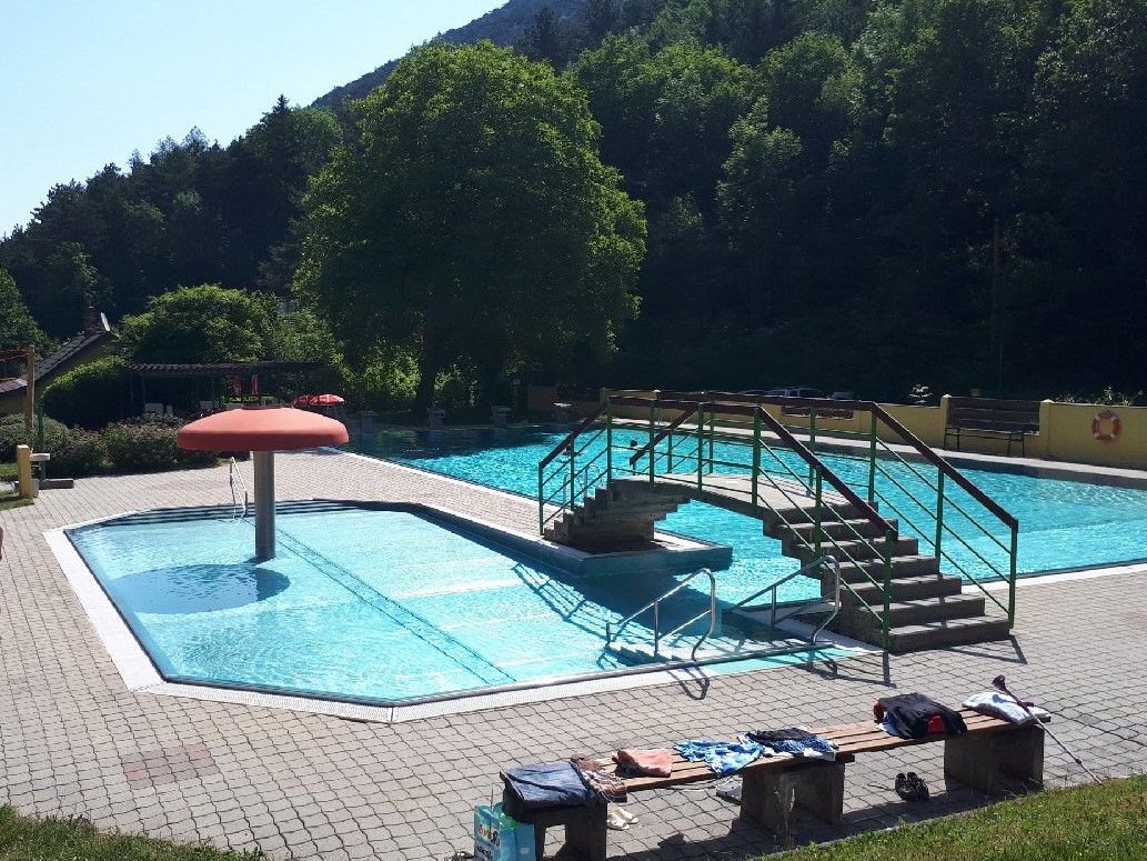 Ein Freibad mit einem kleinen Pool, einer Brücke und einem Pilz-Wasserspiel. Umgeben von Bäumen und einem sonnigen Himmel.