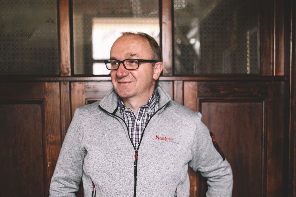 Ein Mann mit Brille und grauer Jacke steht vor einer Holzwand.