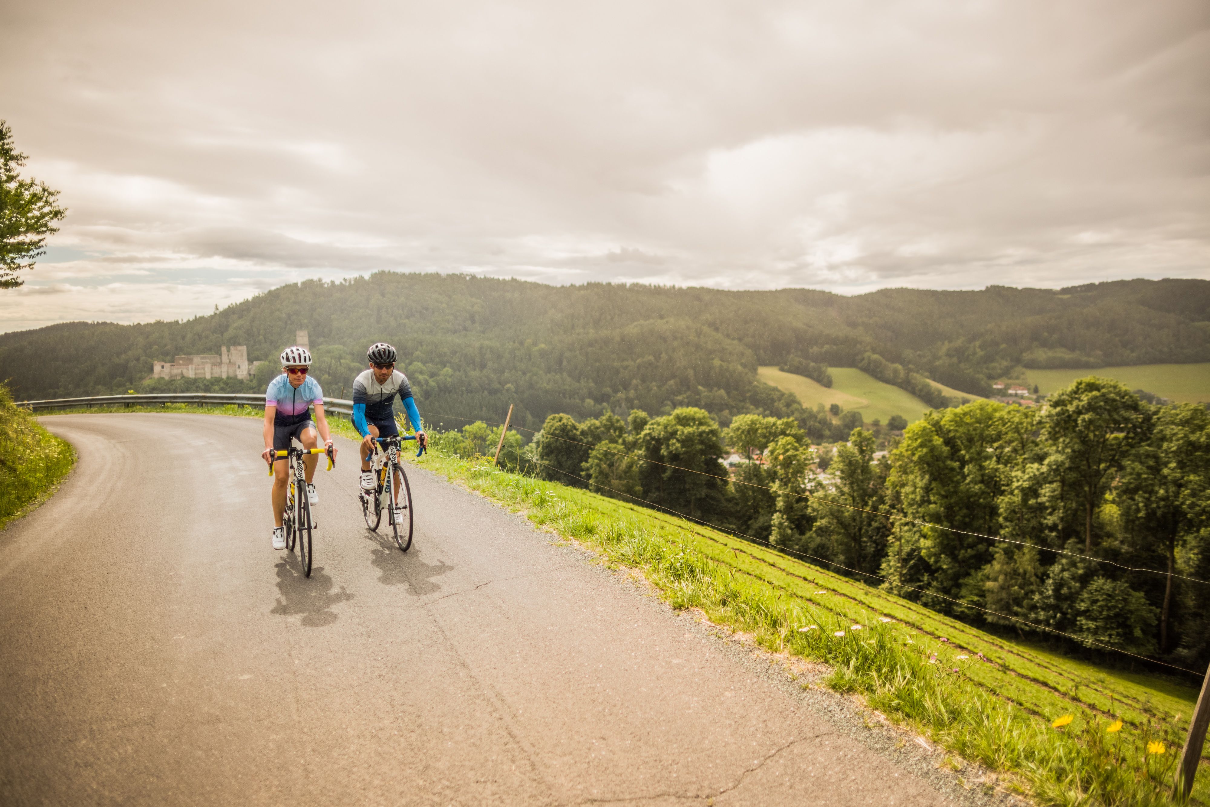 Zwei Radfahrende auf einer kurvigen Straße in einer hügeligen Landschaft mit der Burgruine Kirchschlag im Hintergrund.