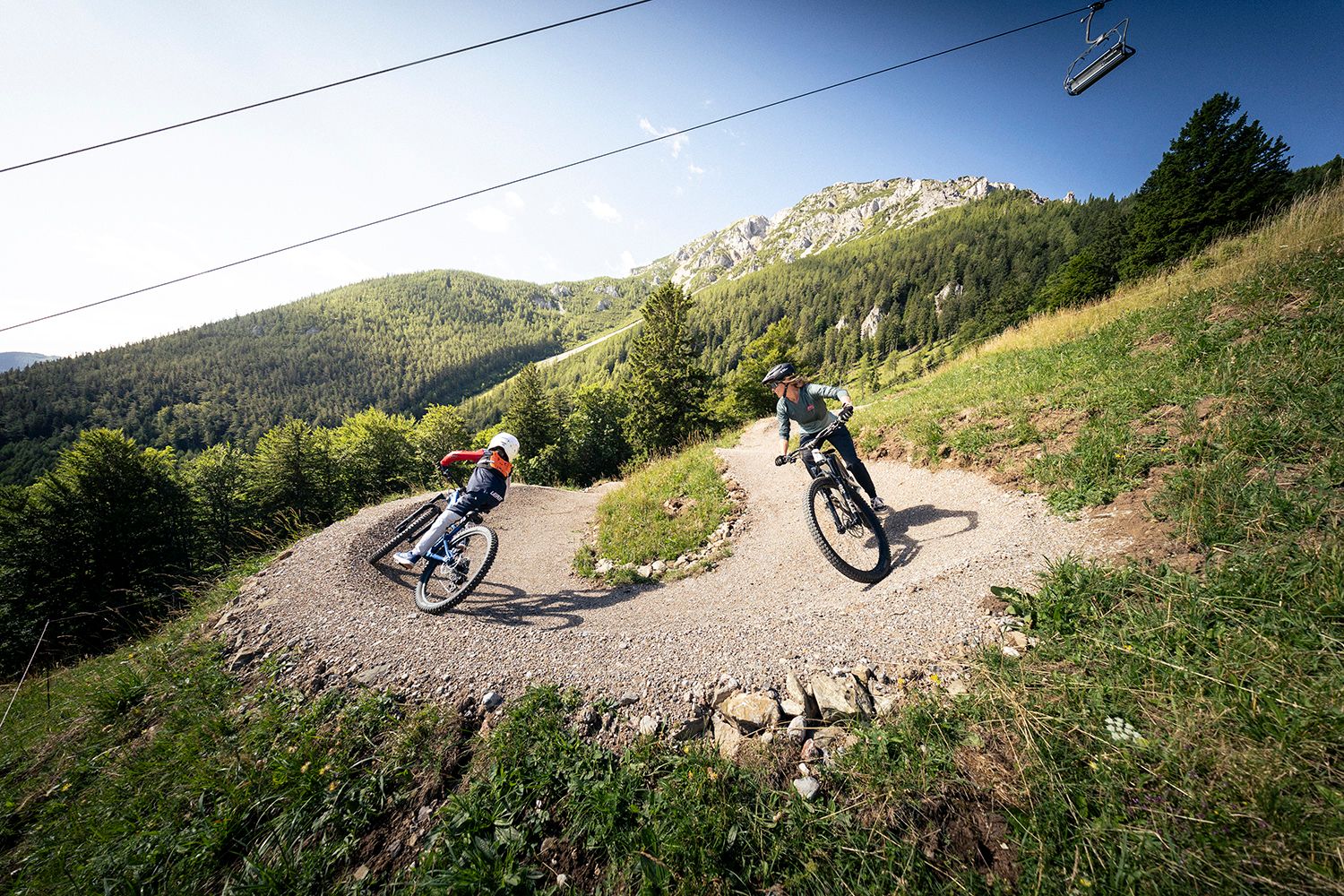 Zwei Mountainbiker fahren auf einem kurvigen Pfad im Alpen Bikepark Schneeberg. Im Hintergrund sind grüne Wälder und Berge zu sehen.
