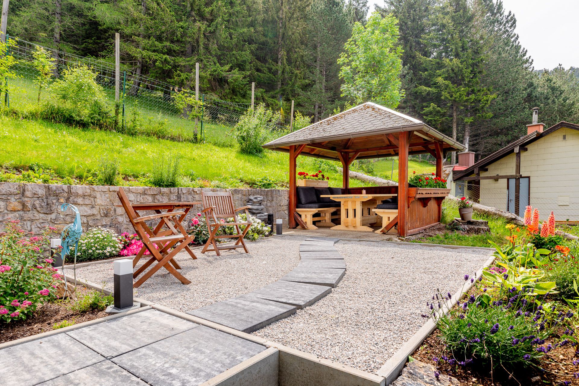 Garten mit Pavillon, Holzmöbeln und Blumenbeeten vor einem Wald.