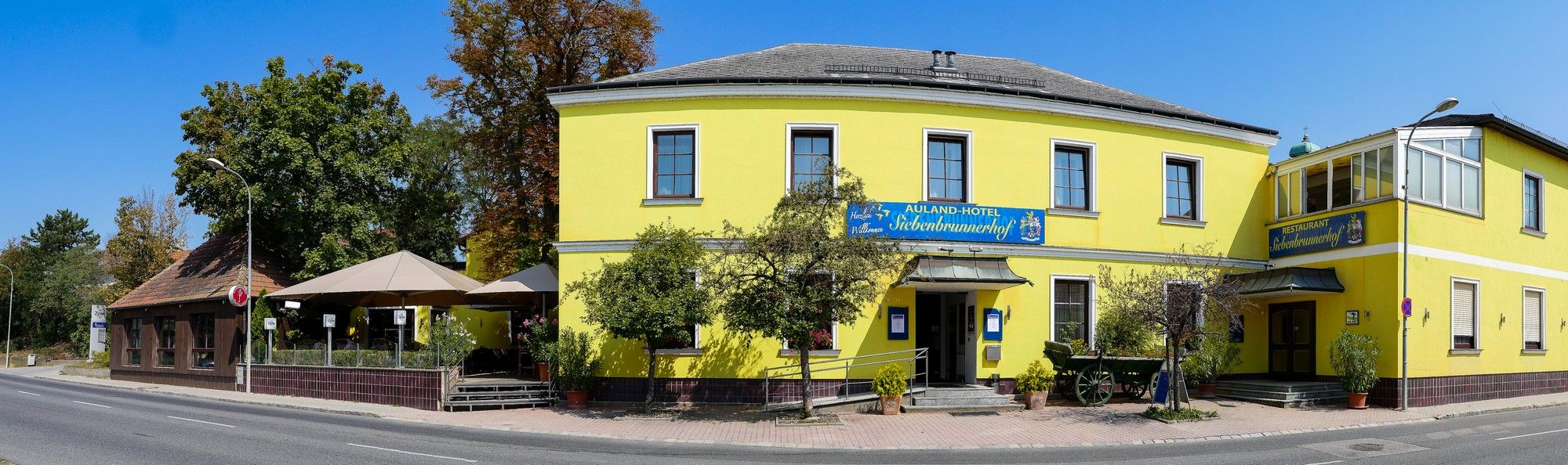 Gelbes Gebäude mit Restaurant und Hotel, umgeben von Bäumen und Straße.