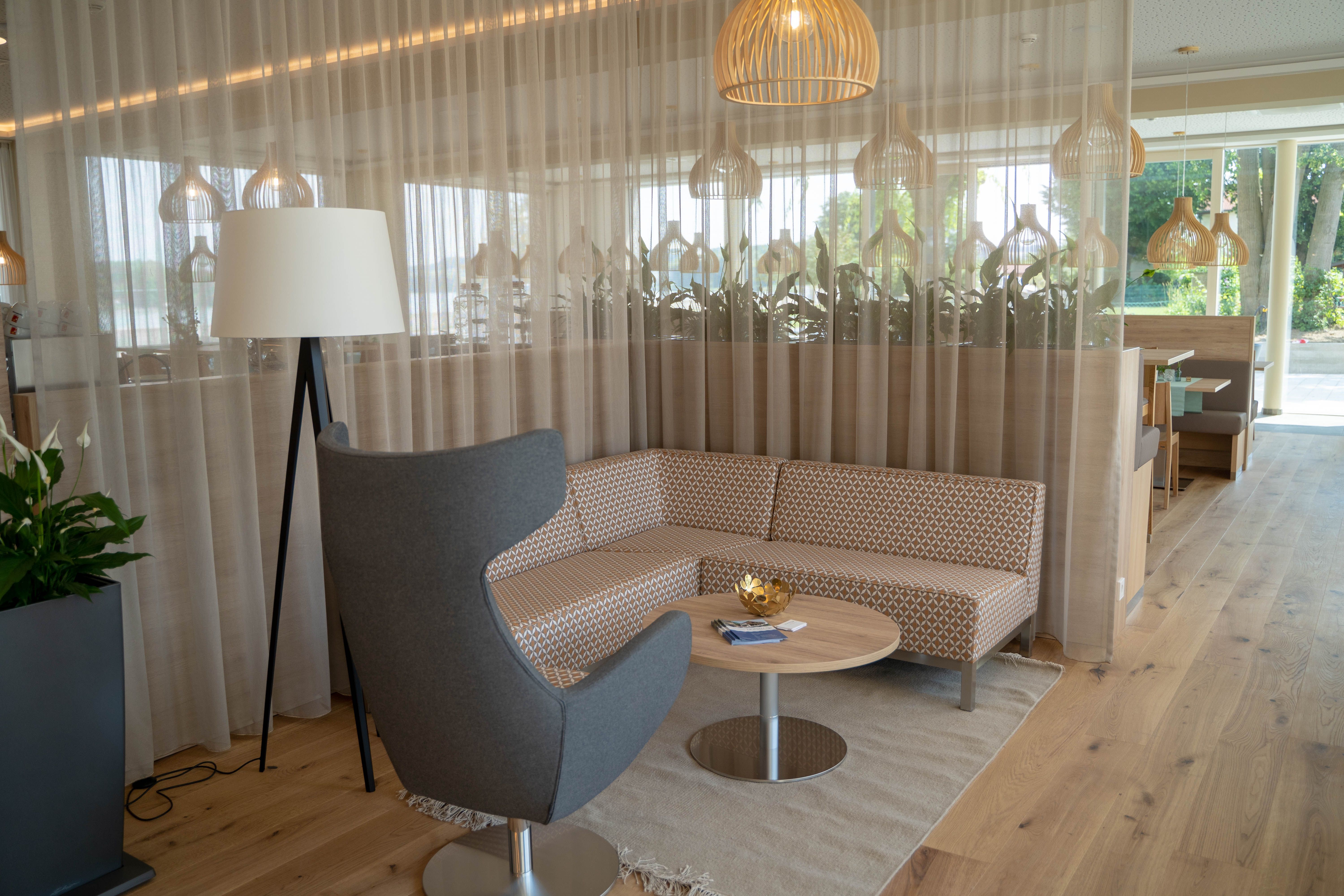 Moderne Sitzecke mit grauem Sessel, gemustertem Sofa, rundem Holztisch und Stehlampe vor einem Vorhang.
