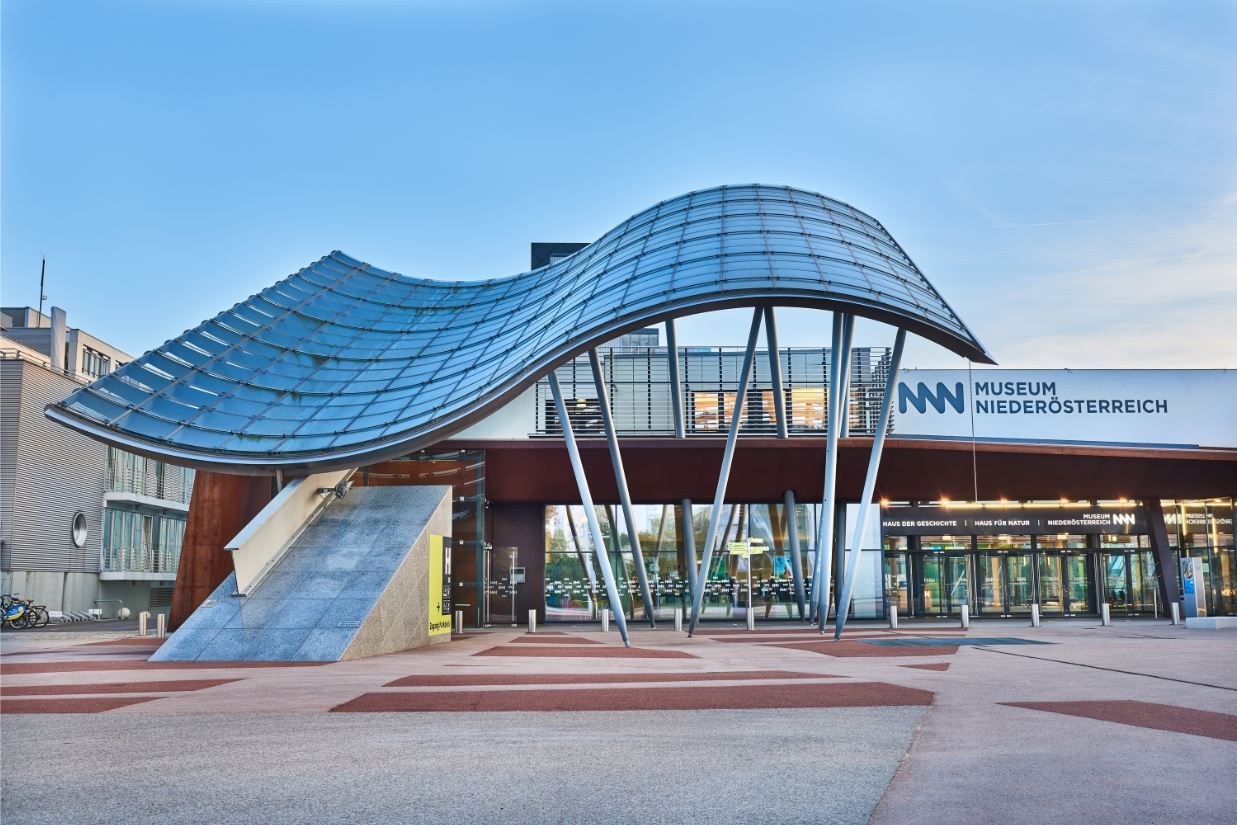 Eingang des Museums Niederösterreich mit moderner Architektur und geschwungenem Glasdach.