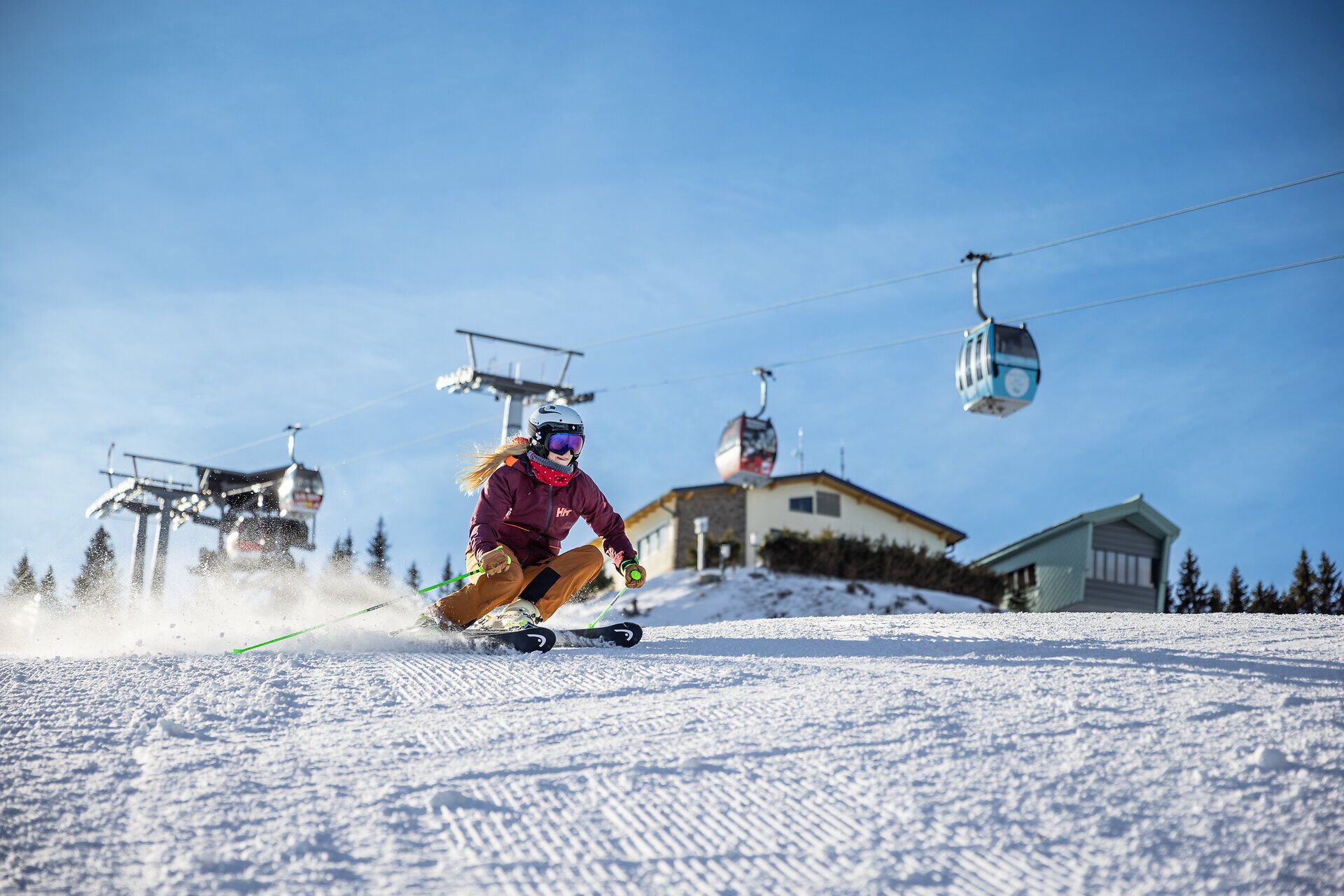 Zwei Wanderer gehen auf einem Weg bergauf, vorbei an einem Aussichtsturm und einem Gebäude, umgeben von Bäumen und einem klaren Himmel.