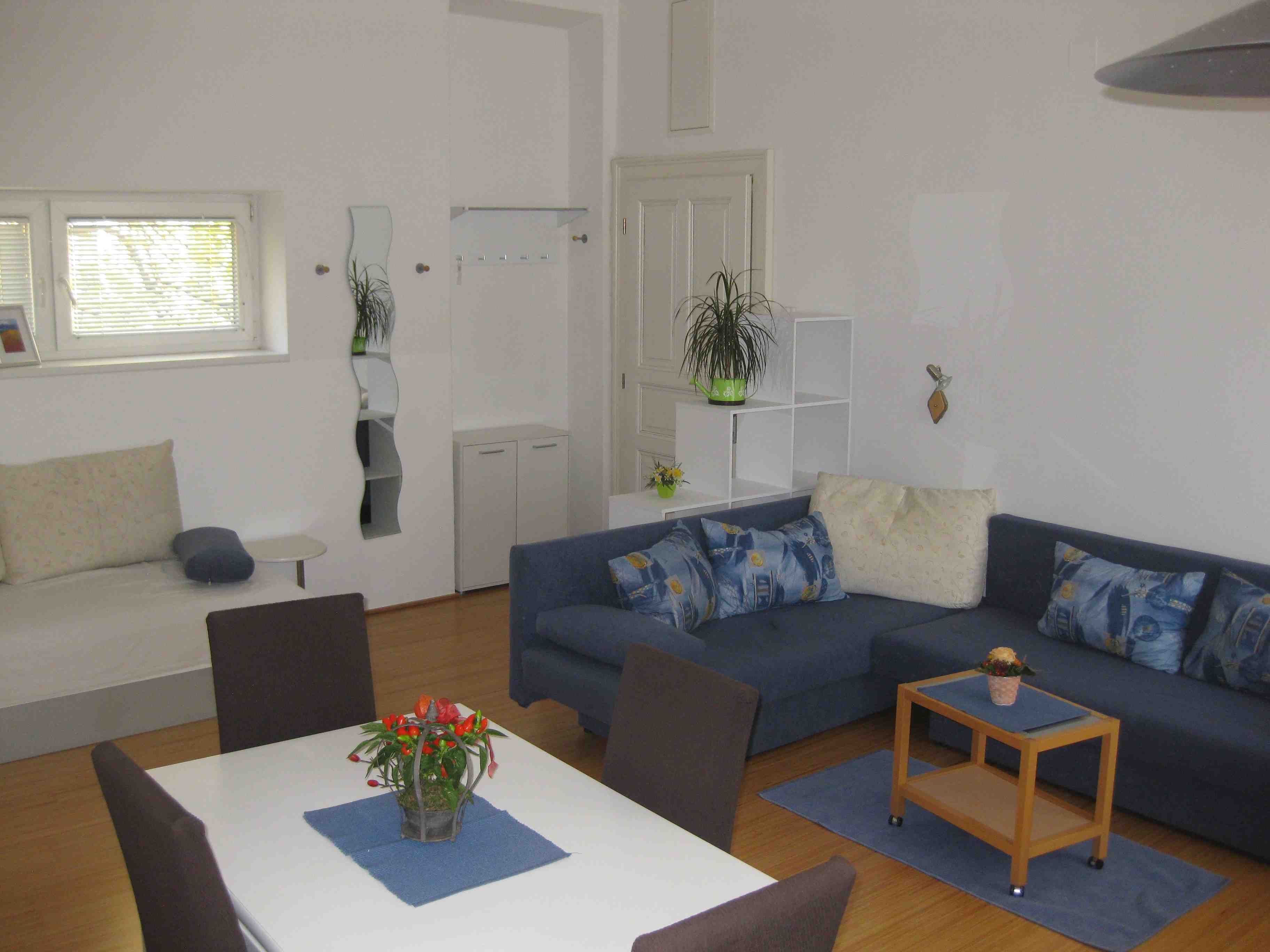 Apartment mit blauen Sofa, weisem Tisch und blauen Stühlen. Im Hintergrund ein kleines Fenster, Tür und Spiegel.