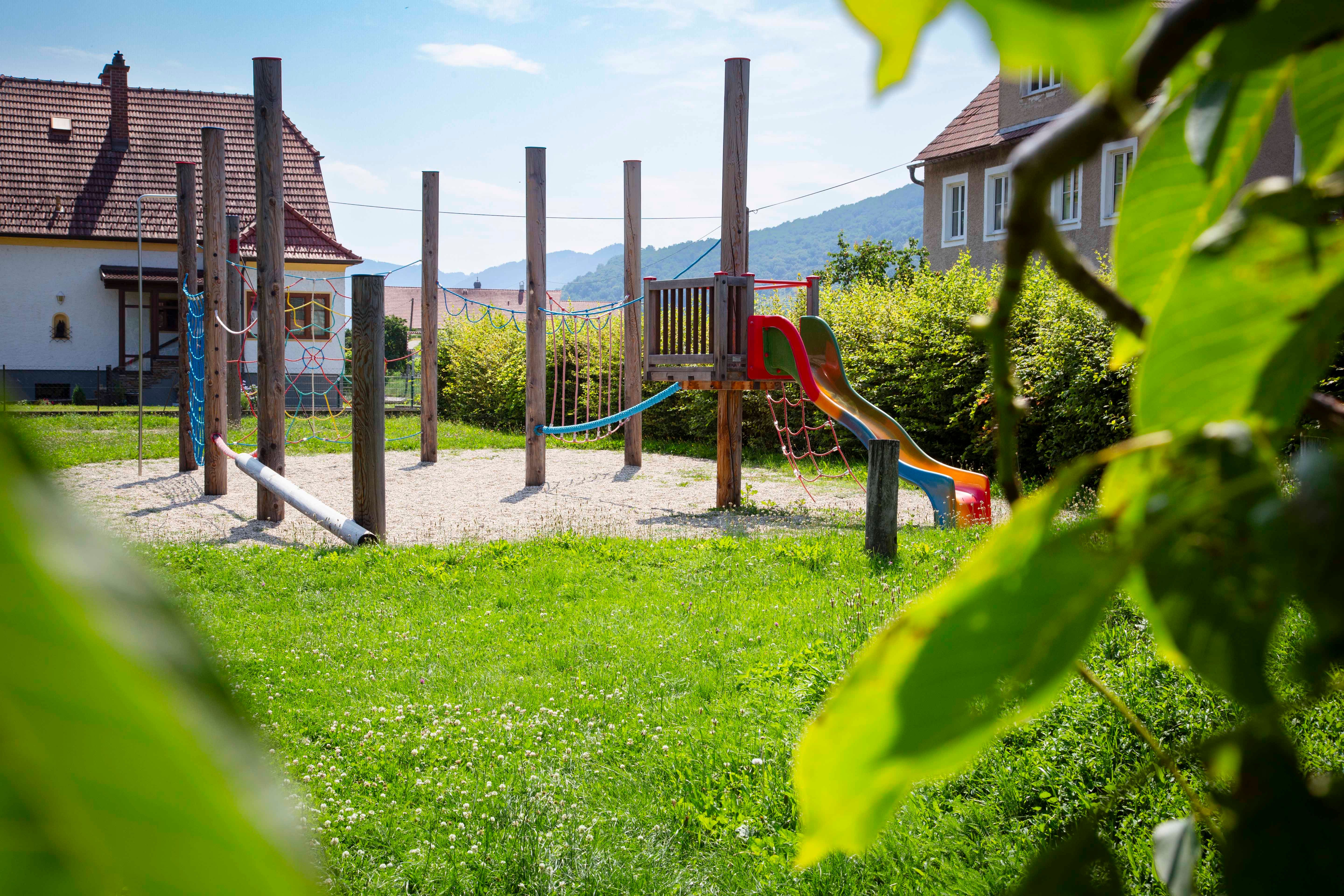 Ein Abenteuerspielplatz mit Klettergerüst und Rutsche, umgeben von grüner Wiese und Bäumen, im Hintergrund Häuser und Hügel.