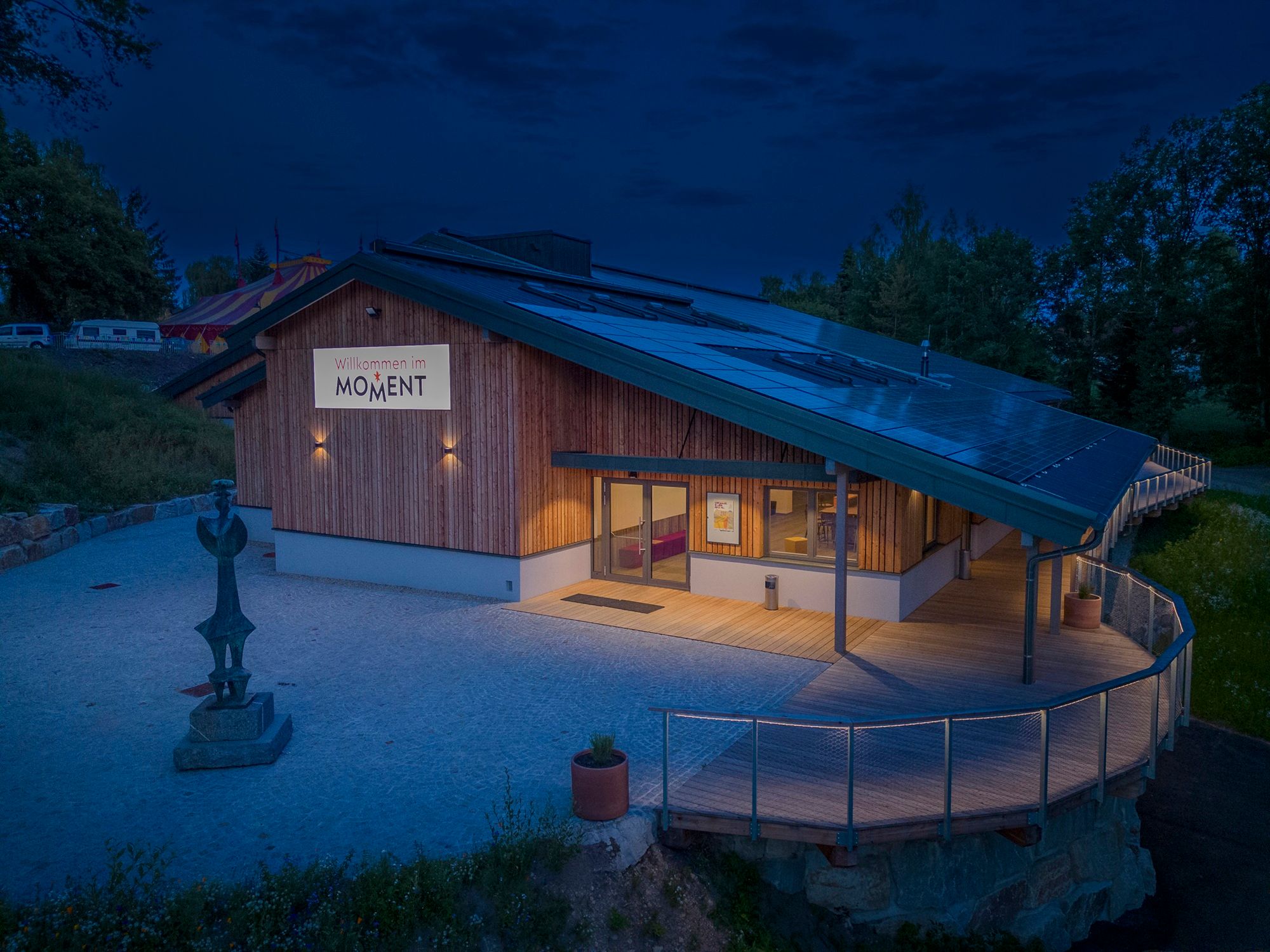 Ein modernes Gebäude mit Holzverkleidung und Solarpanelen auf dem Dach, beleuchtet in der Abenddämmerung. Ein Schild mit der Aufschrift 'Willkommen im MOMENT' ist sichtbar.