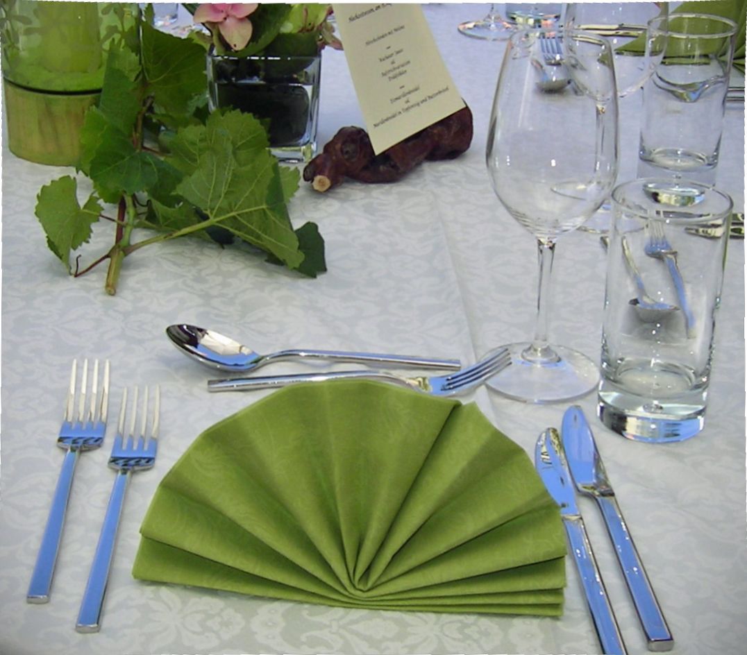 Elegante Tischdekoration mit grün gefalteter Serviette, Besteck, Weinglas und Wasserglas auf weißer Tischdecke.
