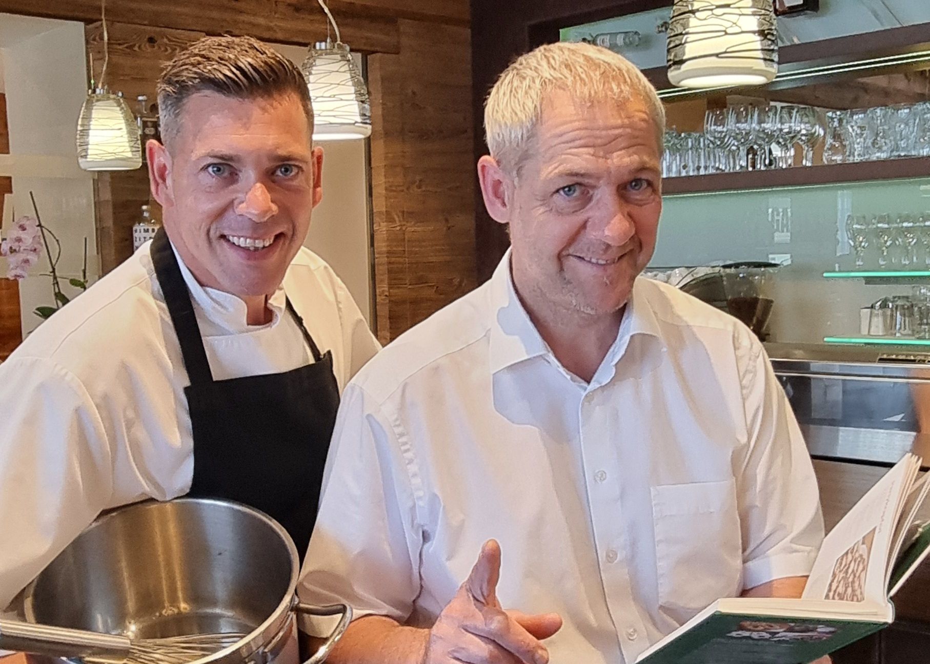 Zwei Männer in einem Restaurant, einer in Kochkleidung mit einem Topf, der andere in einem weißen Hemd mit einem Buch.