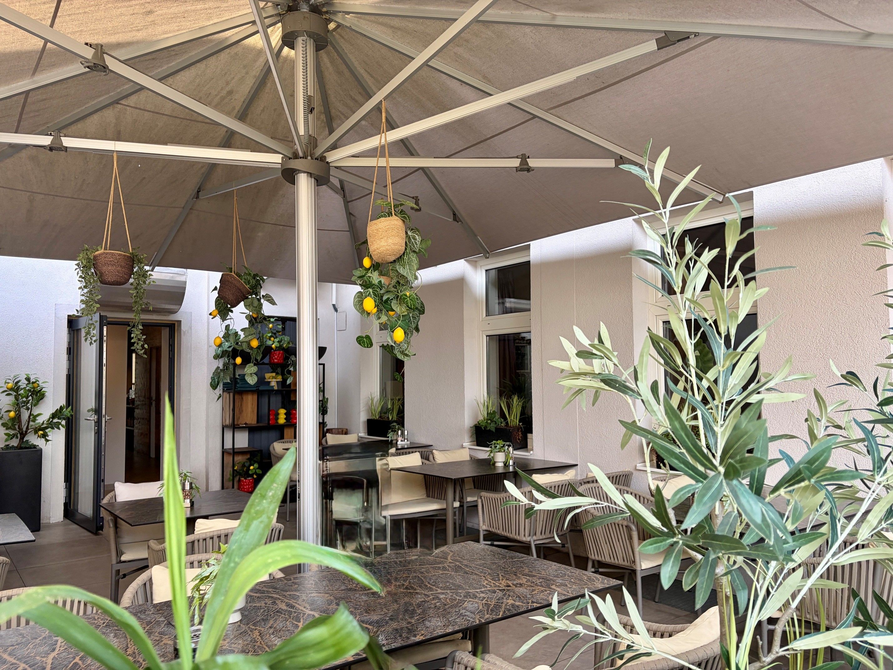 Ein gemütlicher Gastgarten mit Sonnenschirm, hängenden Pflanzen und Tischen, umgeben von grünen Pflanzen und Dekorationen.