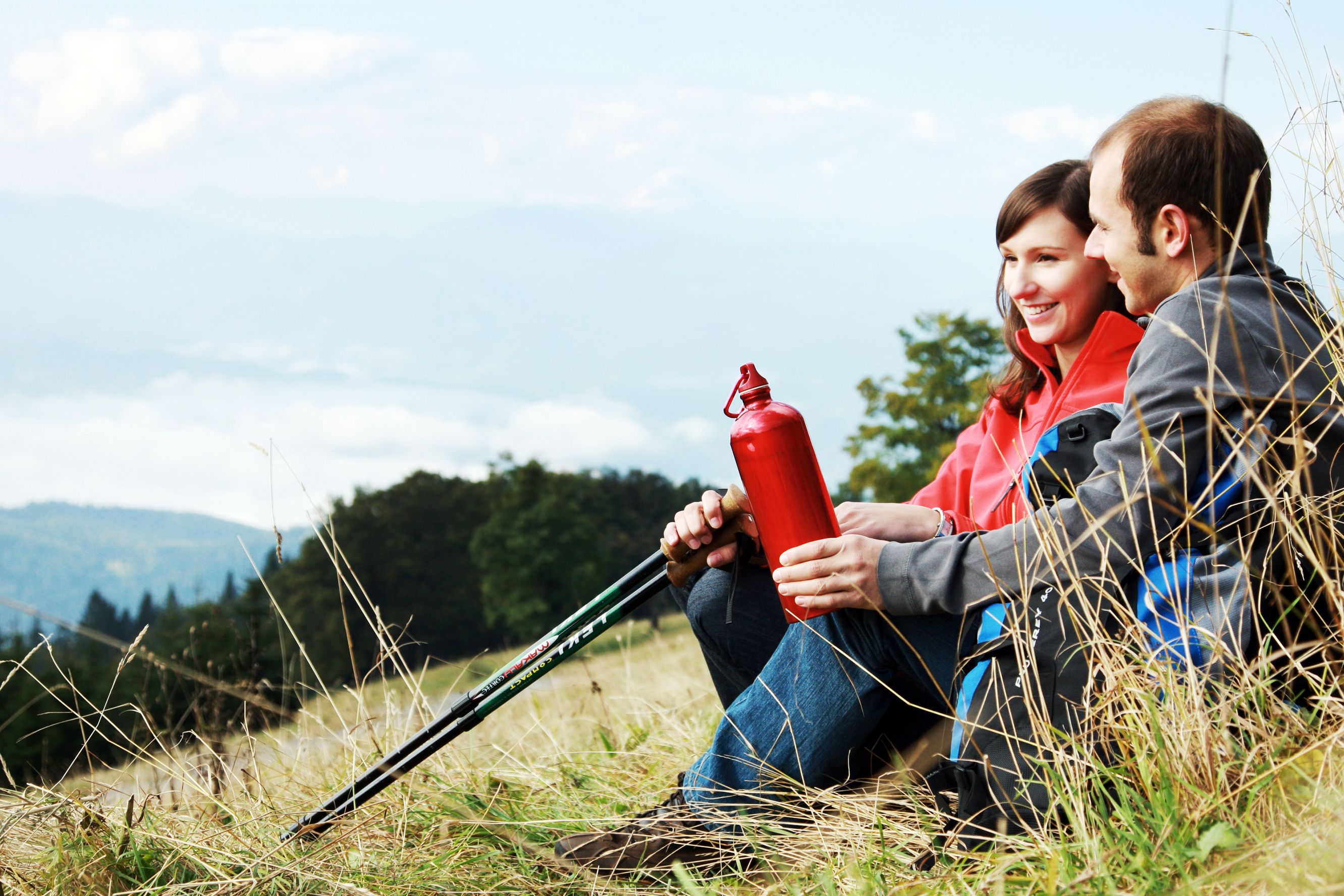 Ein Paar sitzt auf einer Wiese mit Wanderstöcken und einer roten Trinkflasche, im Hintergrund Berge und Wolken.