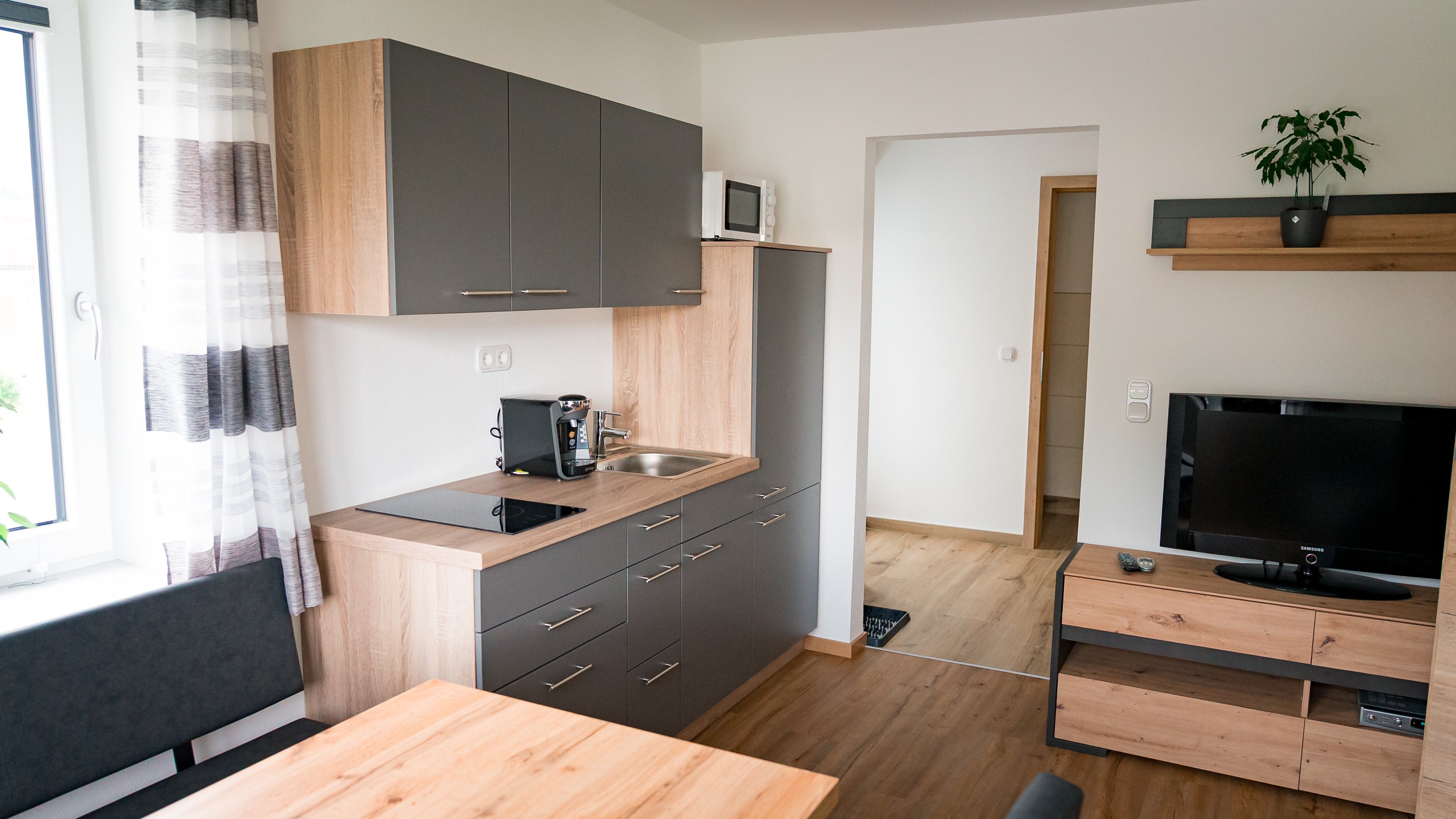 Moderne Küche mit grauen Schränken, Holzarbeitsplatte, Kaffeemaschine, Spüle, Mikrowelle und Fernseher auf Holzregal.
