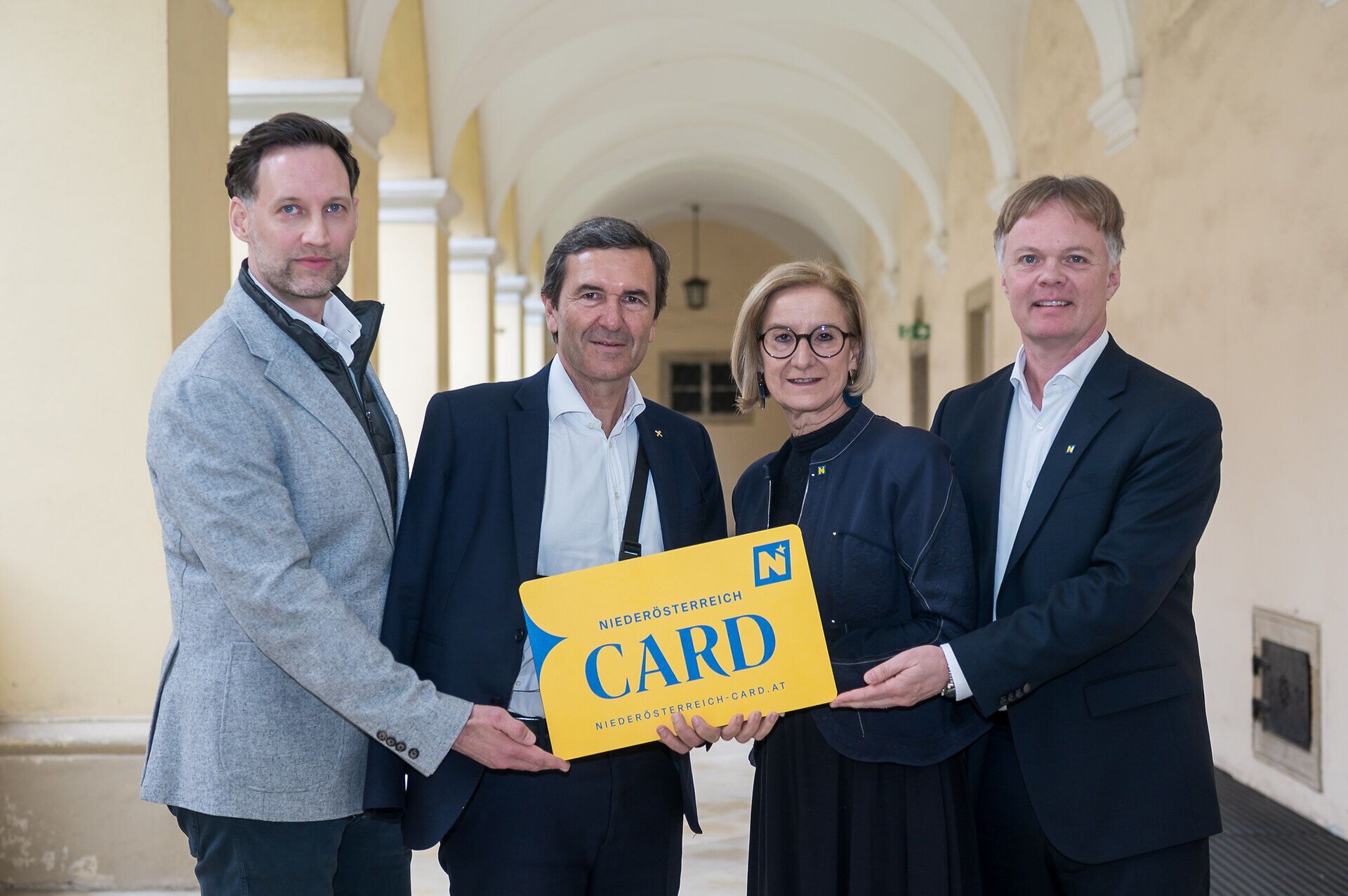 Klemens Wögerer, Reinhard Karl, Landeshauptfrau Johanna Mikl-Leitner und Michael Duscher stehen bei der Pressekonferenz zum Saisonstart 2026 der Niederösterreich-CARD in einem Arkadengang und präsentieren gemeinsam eine große gelbe Niederösterreich-CARD.