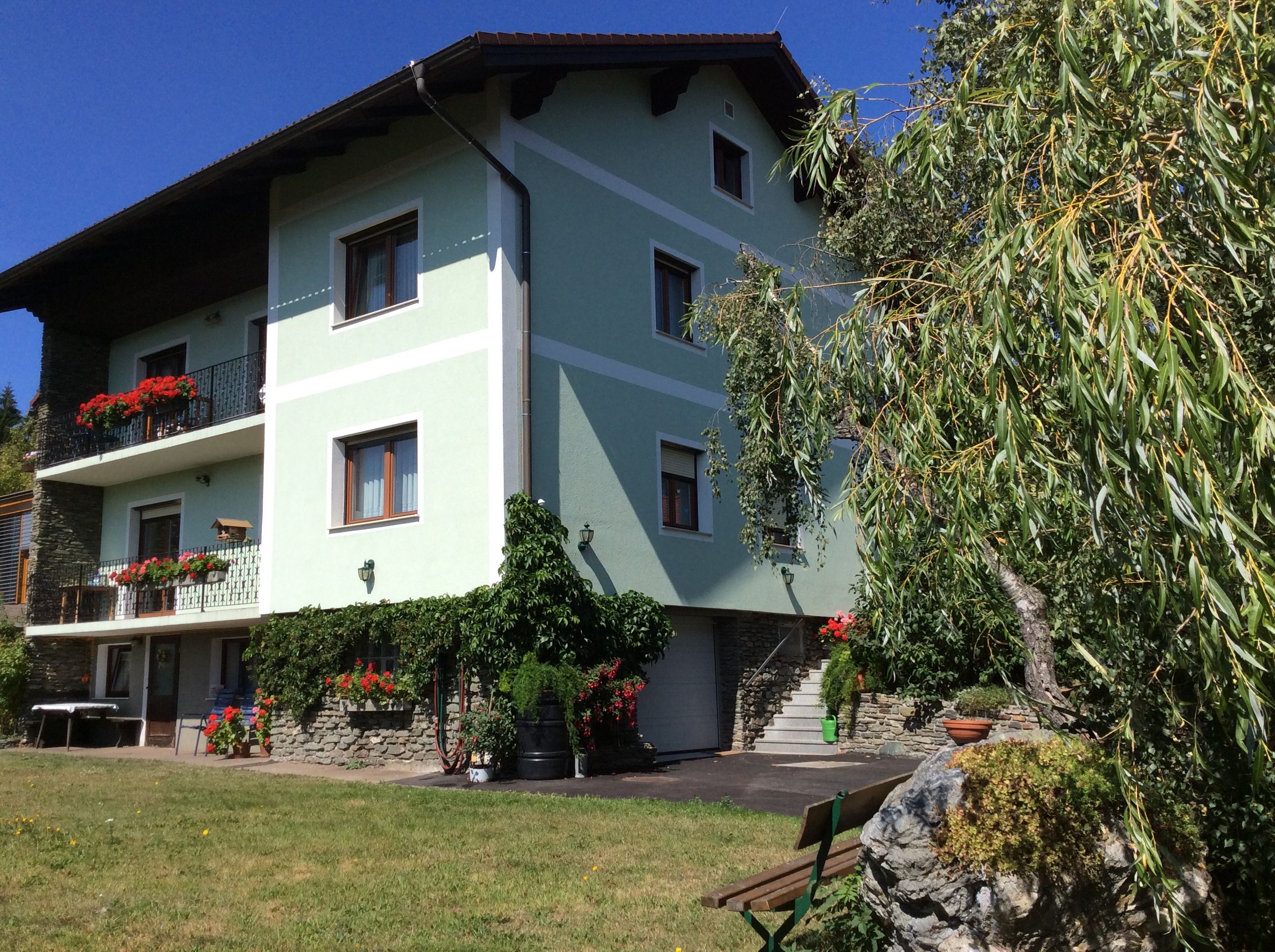 Ein grünes Haus mit Balkonen voller Blumen, umgeben von Bäumen und einem gepflegten Garten.