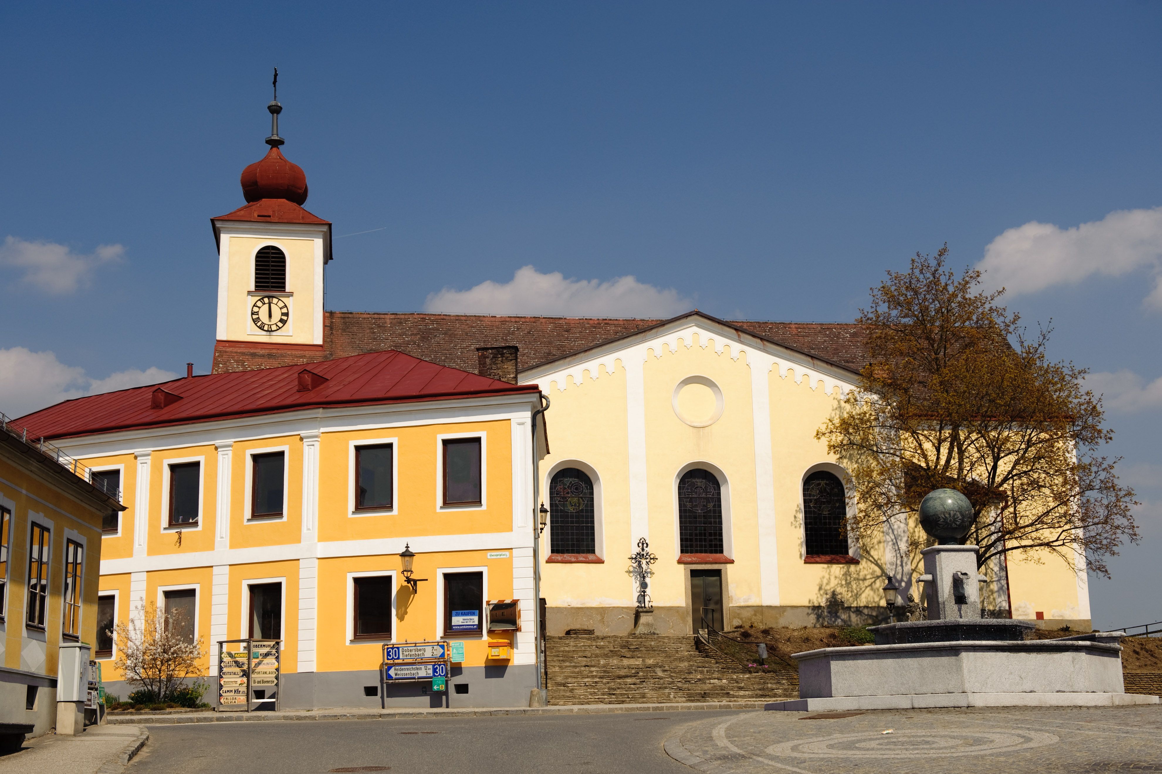 Kirche mit gelber Fassade und Turm mit rotem Dach auf einem Hauptplatz.