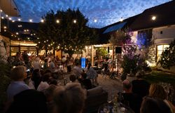 Abendliches Konzert in einem stimmungsvoll beleuchteten Innenhof: Zahlreiche Gäste sitzen an Tischen und verfolgen den Auftritt eines Musikers.