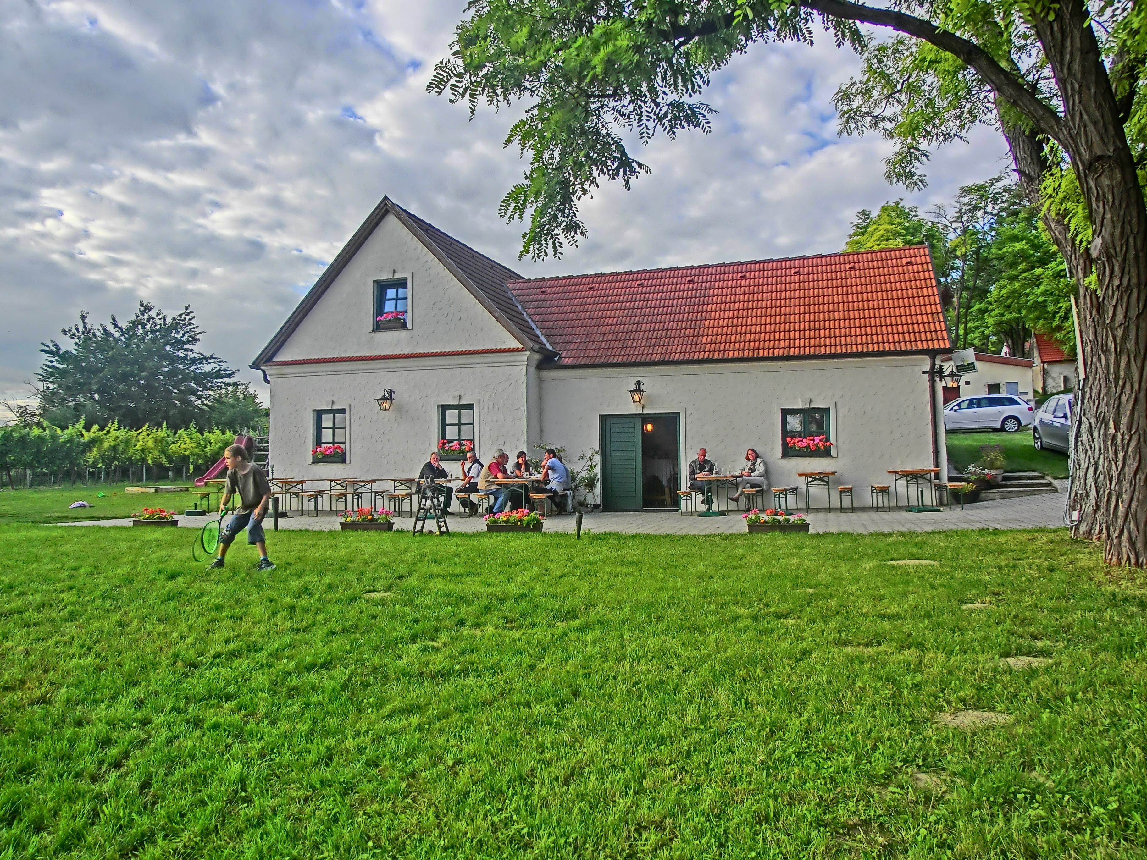 Ein traditionelles Gebäude mit rotem Dach, umgeben von grüner Wiese und Bäumen, Menschen sitzen an Tischen im Freien.