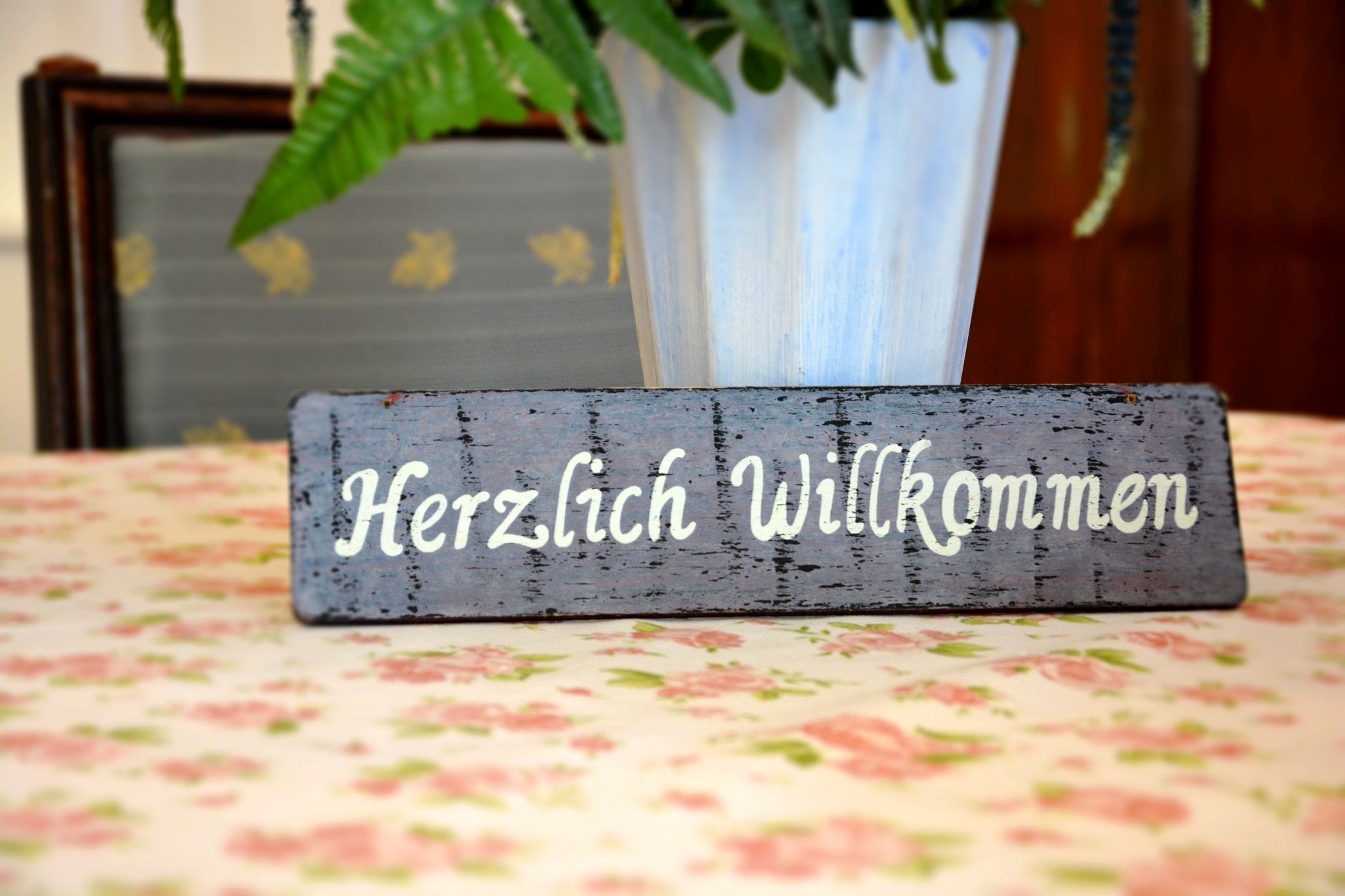 Ein Holzschild mit der Aufschrift 'Herzlich Willkommen' steht auf einem blumigen Tisch.