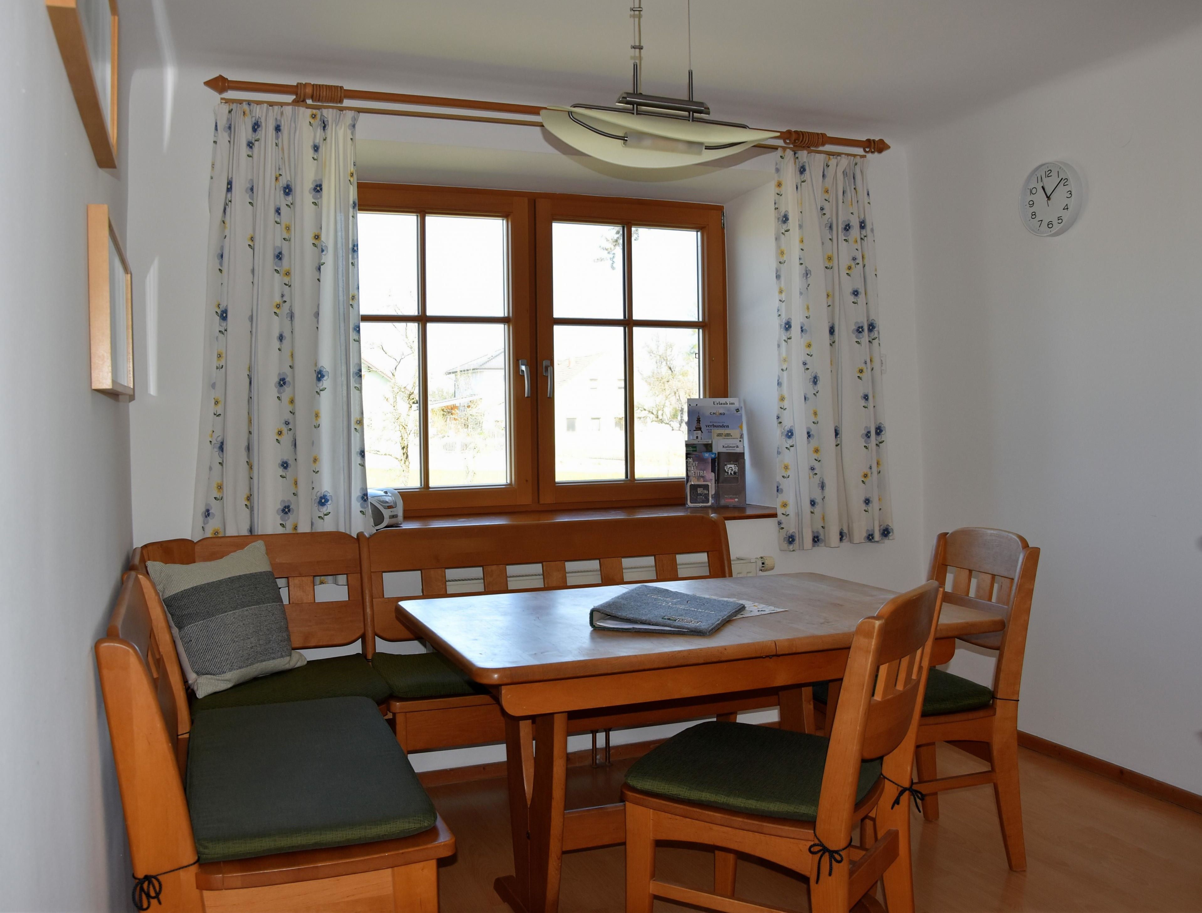 Esszimmer mit Holztisch, vier Stühlen, Eckbank, Fenster mit Vorhängen und Wanduhr.