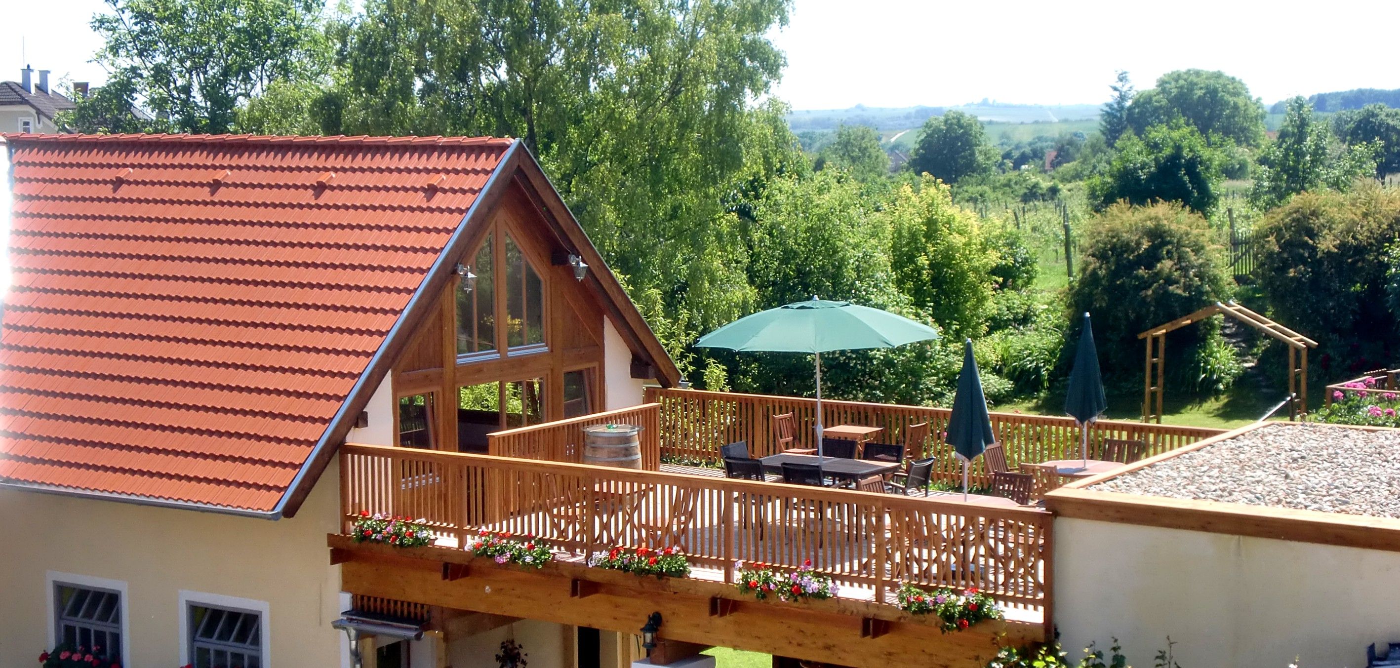 Terrasse mit Holzzaun, Sonnenschirm und Gartenmöbeln, umgeben von grüner Landschaft.