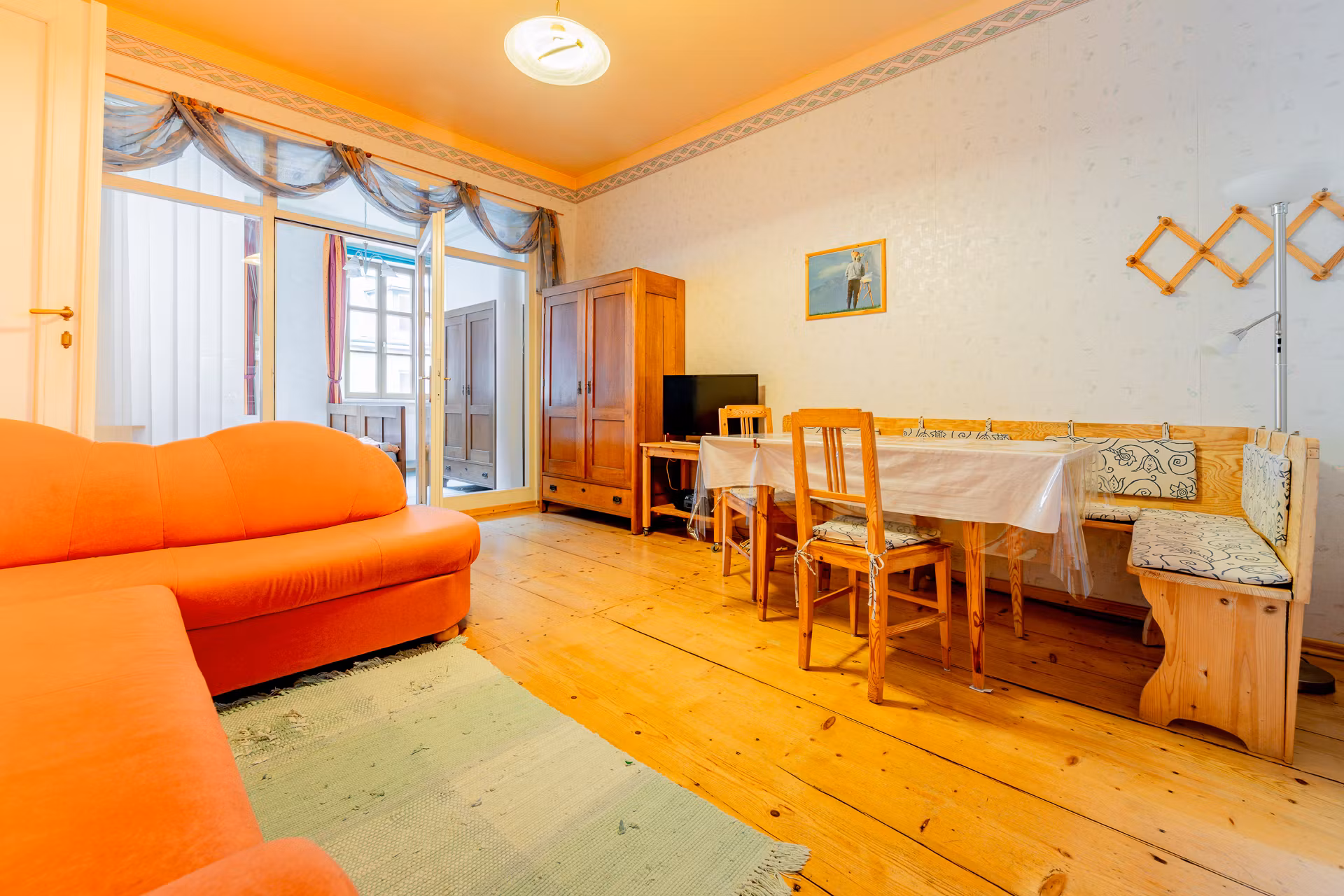 Ein Wohnzimmer mit orangefarbenem Sofa, Holztisch und Stühlen, Holzboden und einem Schrank. Ein Bild hängt an der Wand.