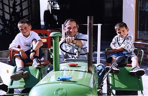 Ein Mann sitzt auf einem grünen Traktor, flankiert von zwei Jungen auf Spielzeugtraktoren.