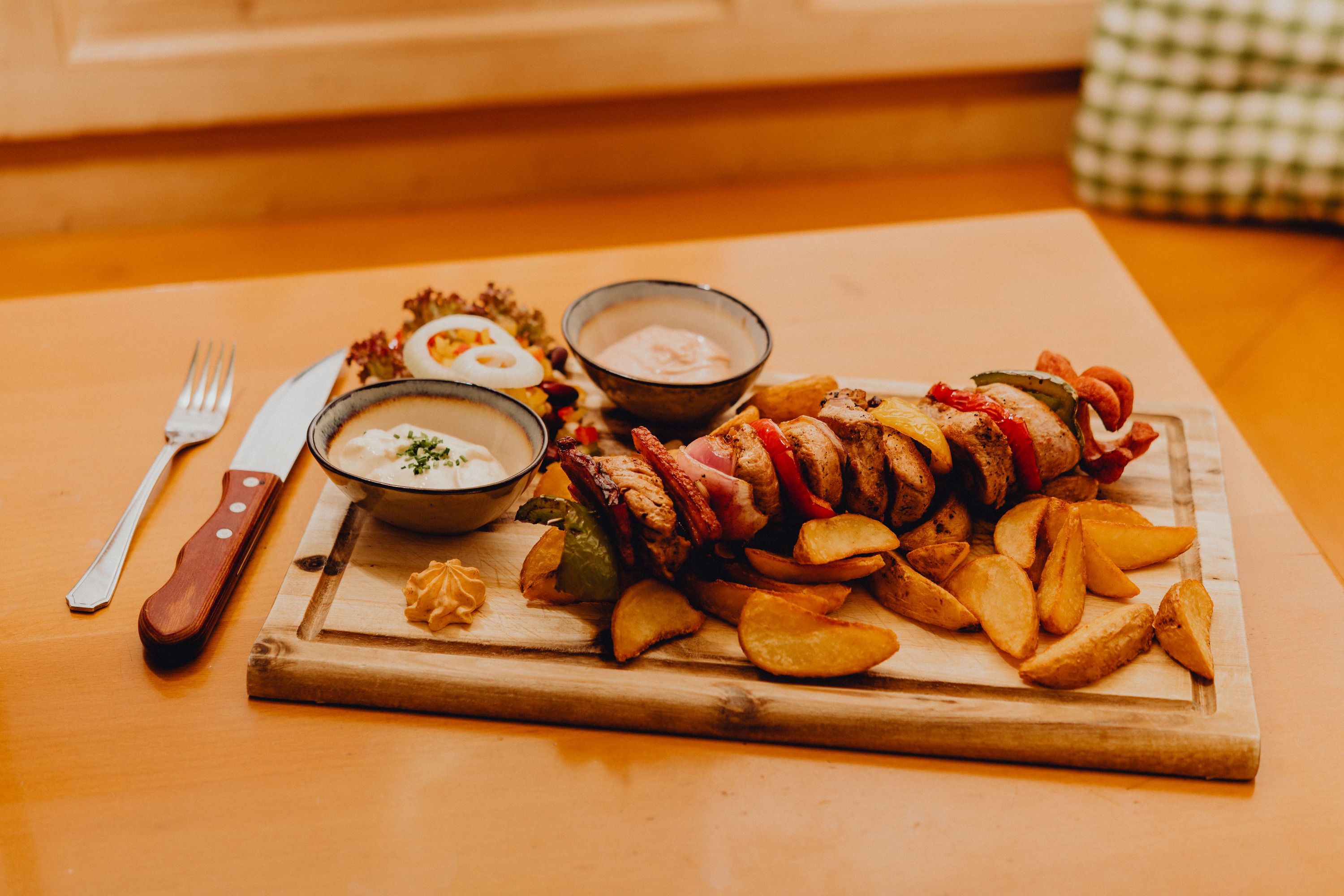 Ein Holzbrett mit einem Fleischspieß, Kartoffelecken, zwei Saucenschalen und Salat auf einem Tisch.
