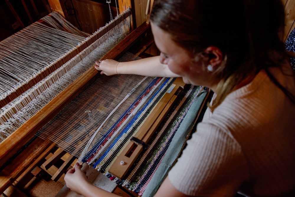 Eine Frau webt an einem Webstuhl einen bunten Fleckerlteppich.