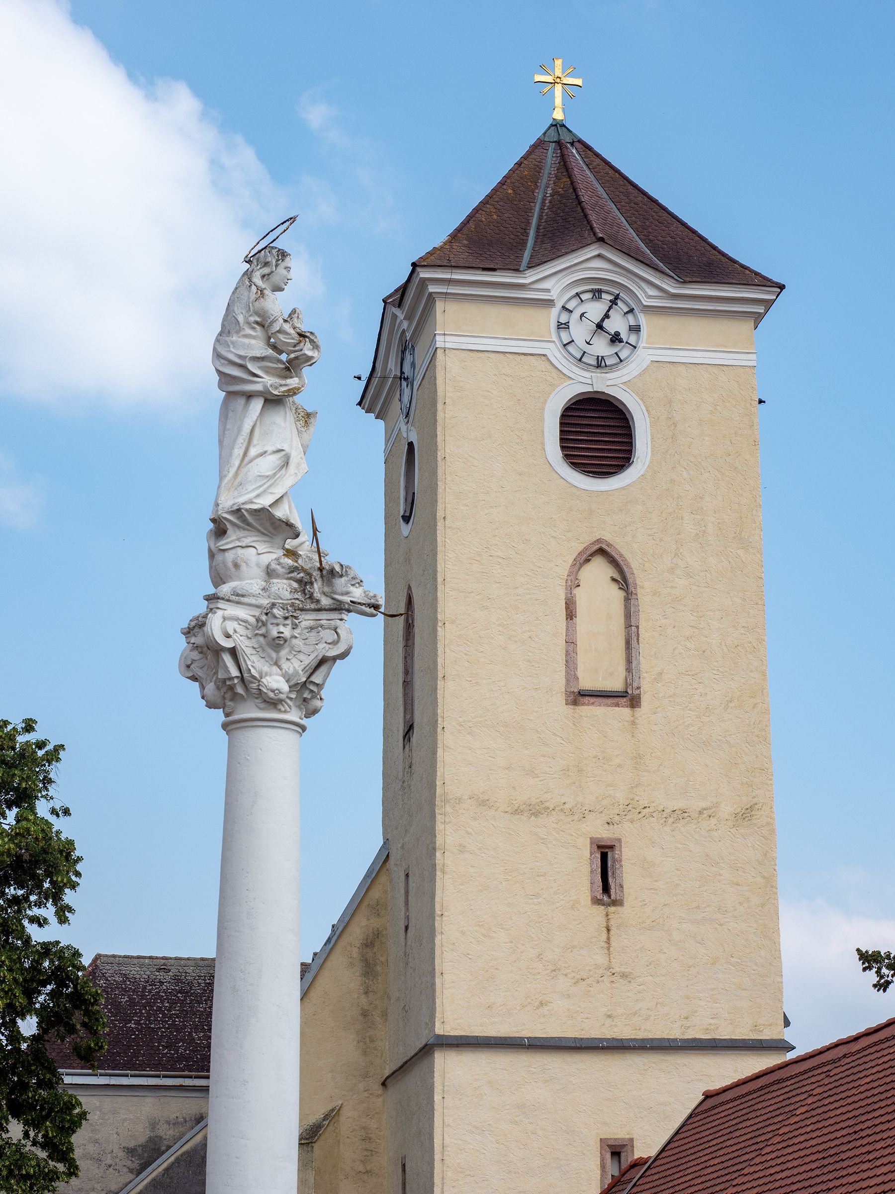 Kirchturm mit Uhr und Mariensäule in Schrattenthal vor blauem Himmel.