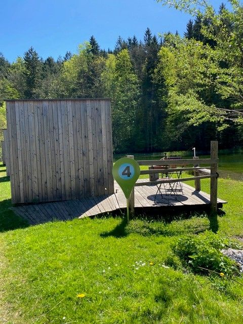 Eine kleine Holzhütte mit Terrasse und Tisch im Grünen, umgeben von Bäumen, direkt am Teich gelegen. 