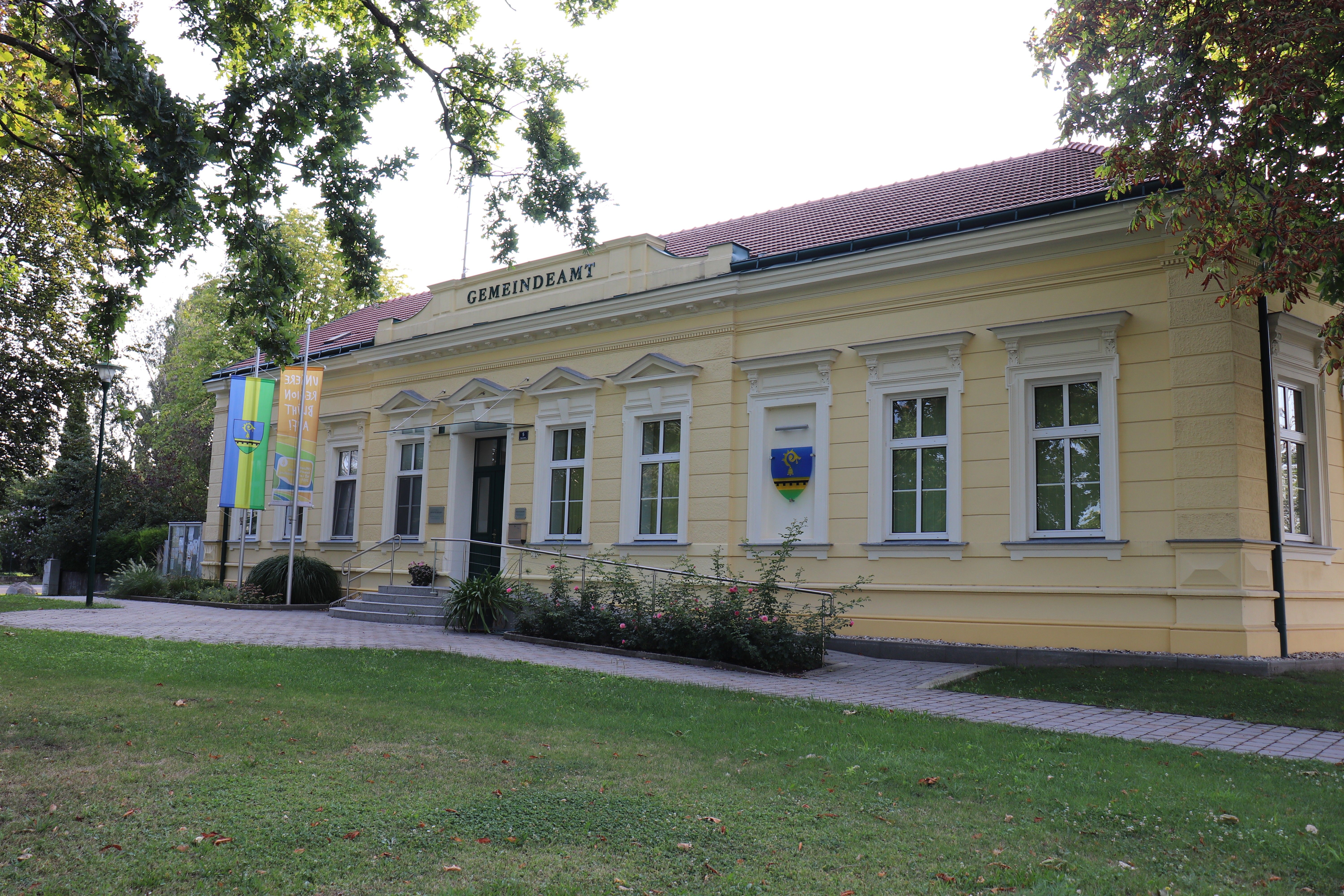 Gemeindeamt Raasdorf, ein helles Verwaltungsgebäude mit rotem Dach, Flaggen und Wappen, umgeben von Bäumen und Rasen.