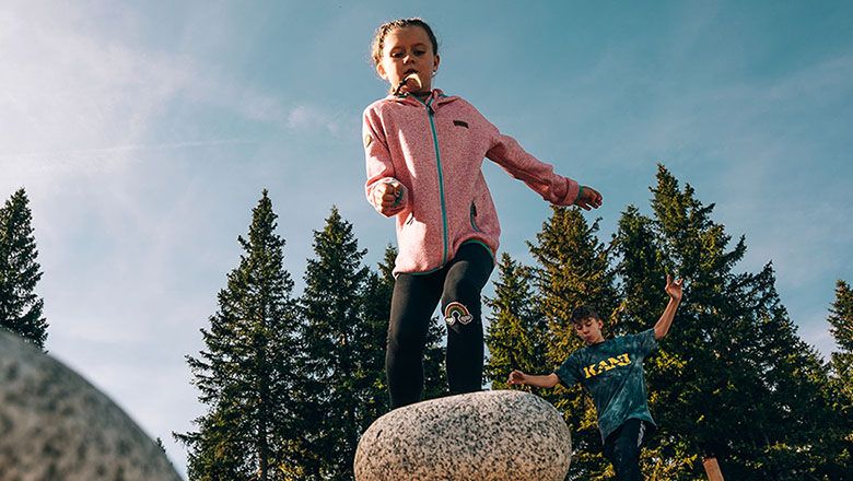 Kinder spielen auf Wackelsteinen vor einem Hintergrund aus Bäumen und blauem Himmel.