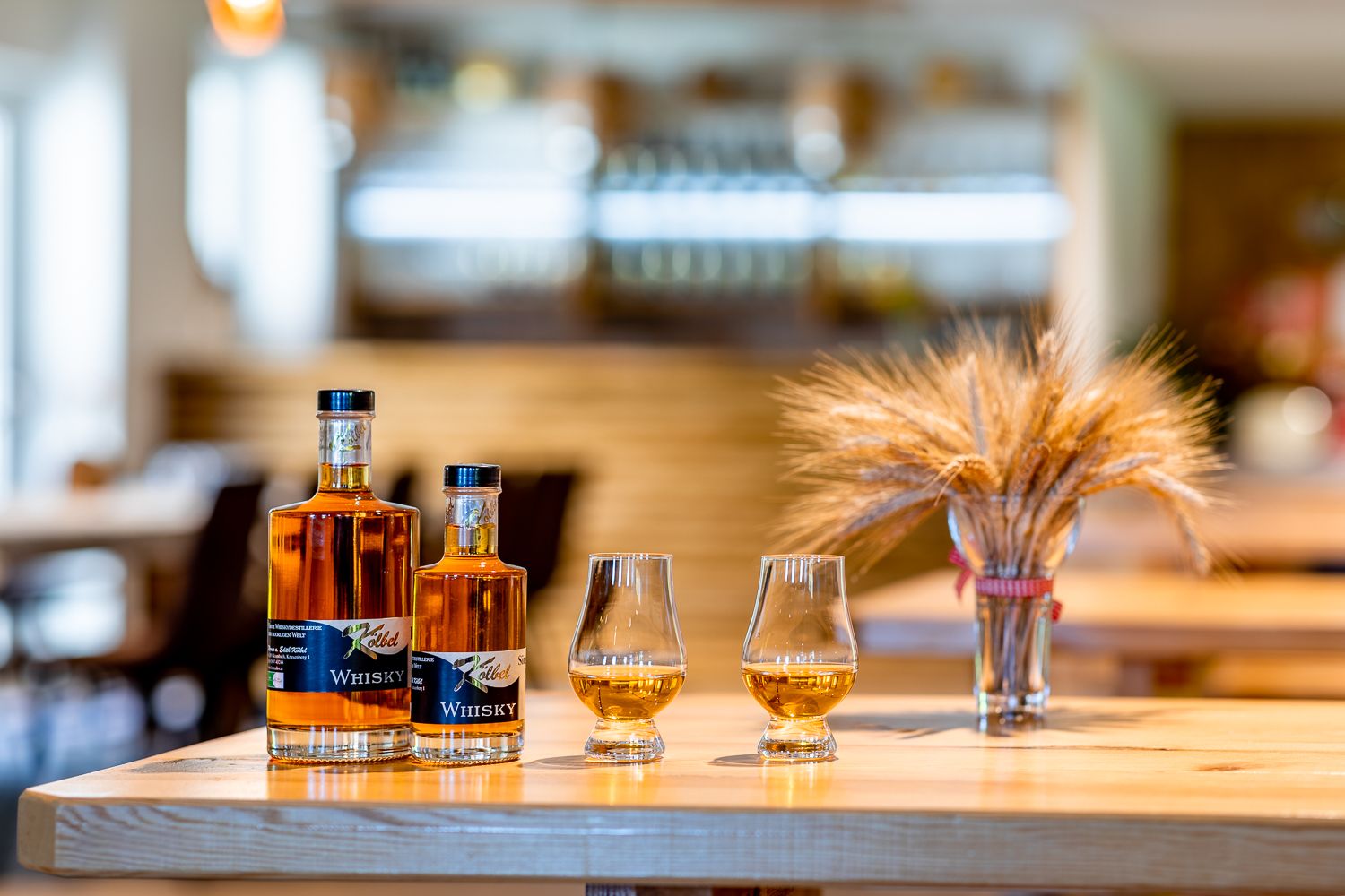 Zwei Whiskyflaschen und zwei Gläser mit Whisky stehen auf einem Holztisch, daneben ein Strauß Getreide.