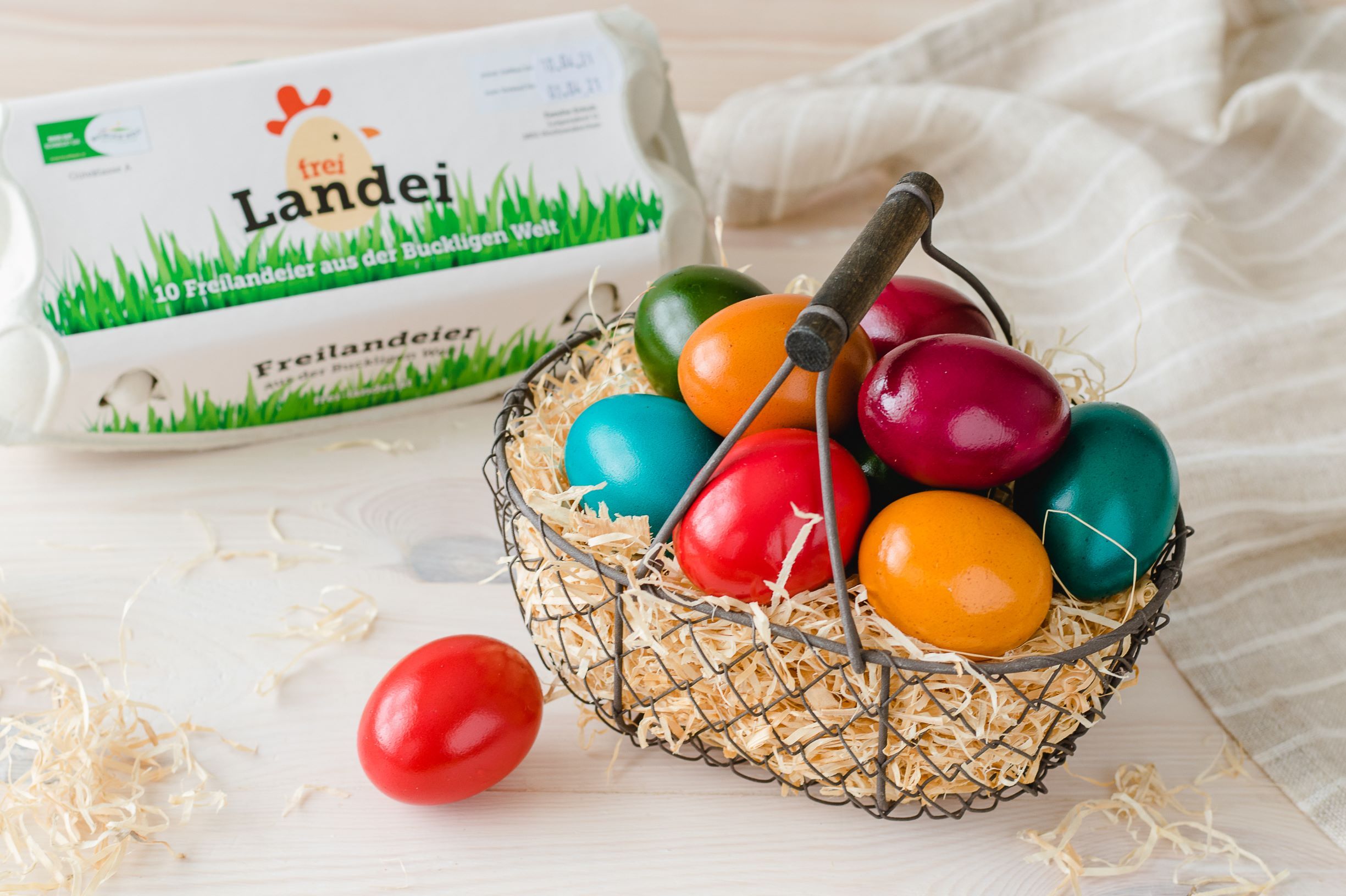 Ein Korb gefüllt mit Stroh und bunten Ostereiern auf einem Holztisch. Daneben ein Eierkarton, ein rotes Ei und ein graues Tuch.