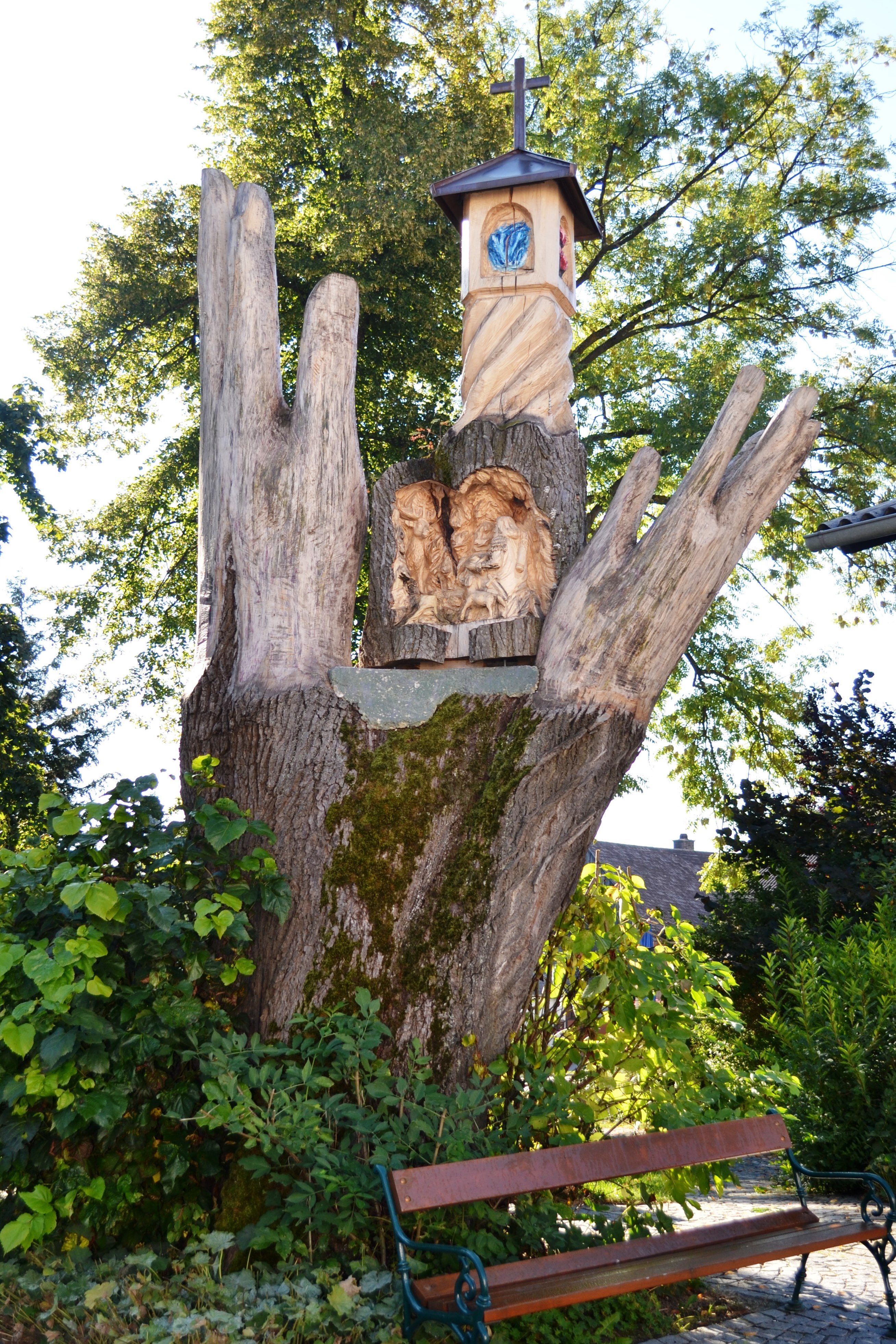 Holzskulptur in Form einer Hand mit geschnitzten Figuren und einem kleinen Turm mit Kreuz.