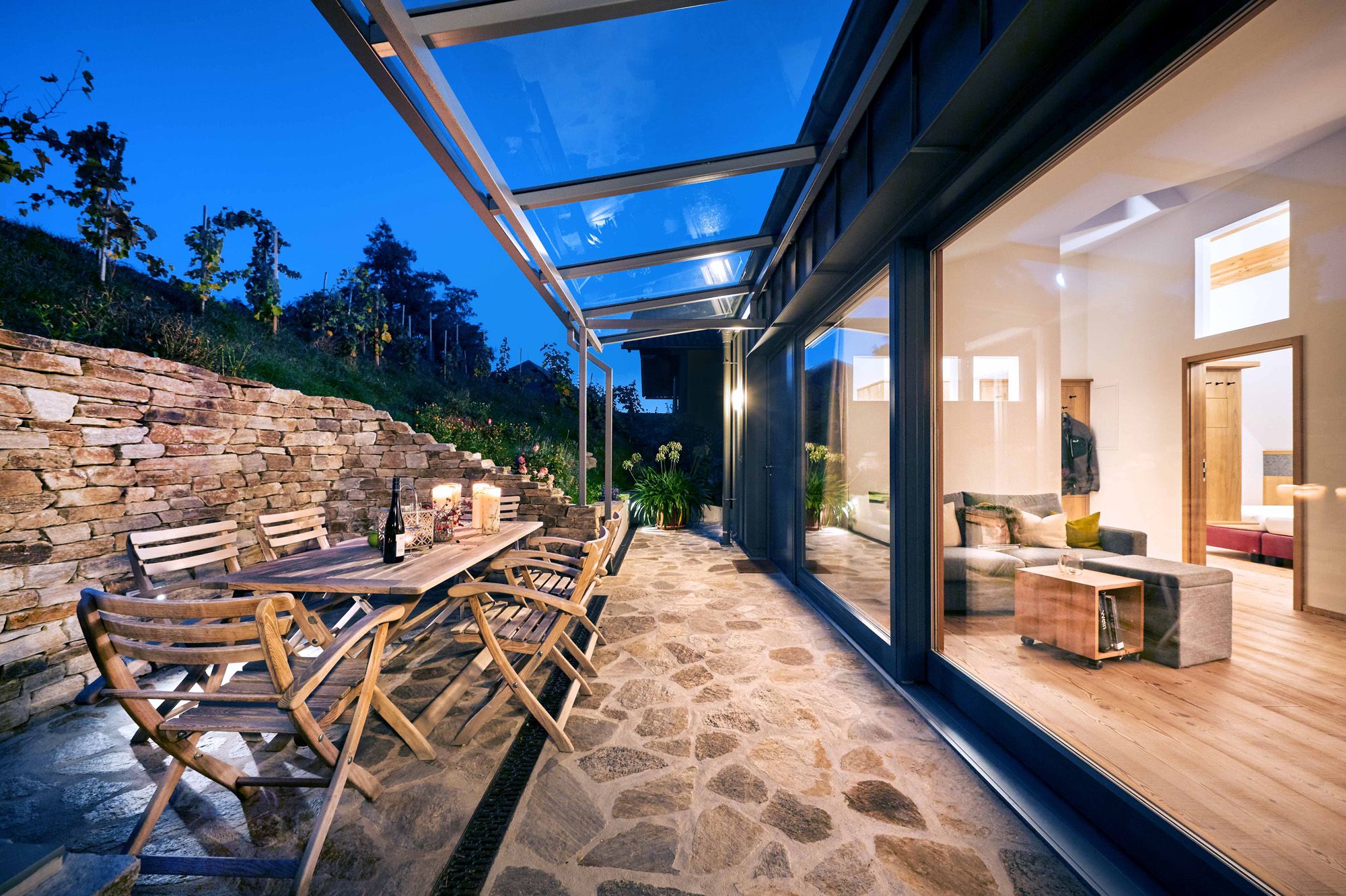 Terrasse mit Holzmöbeln und Glasdach, Blick ins beleuchtete Wohnzimmer.