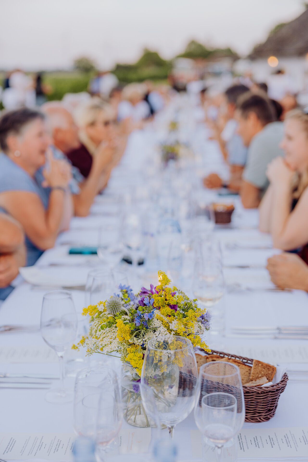 Eine lange Tafel, geschmückt mit bunten Blumen, lädt zu einem geselligen Abendessen ein. Umgeben von der malerischen Kellergasse „Alte Geringen“ genießen die Gäste die kulinarischen Köstlichkeiten der Region. Die Atmosphäre ist geprägt von fröhlichem Lachen und dem Duft von frisch zubereiteten Speisen.