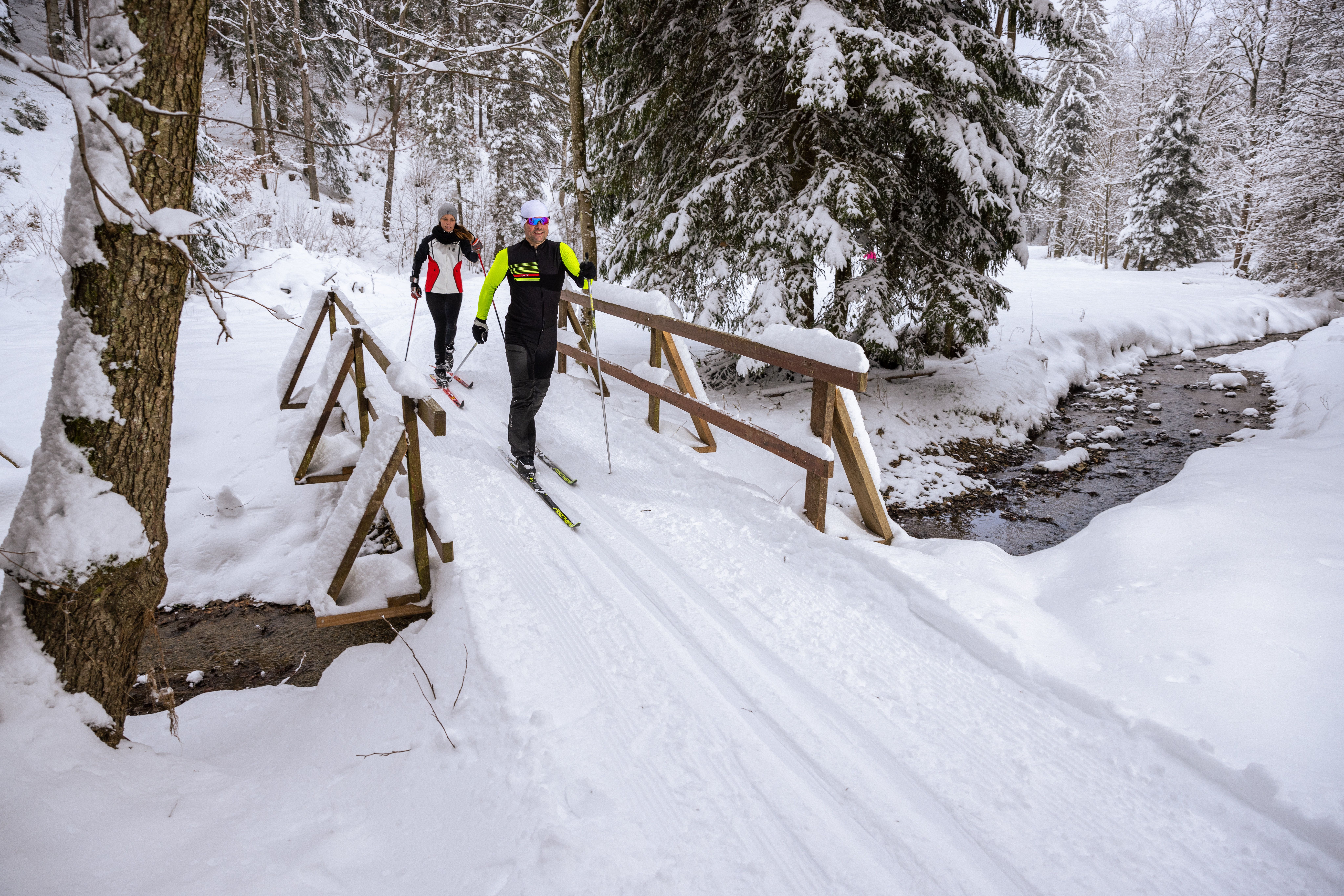 Zwei Personen überqueren beim Langlaufen einen Bach in einer verschneiten Waldlandschaft