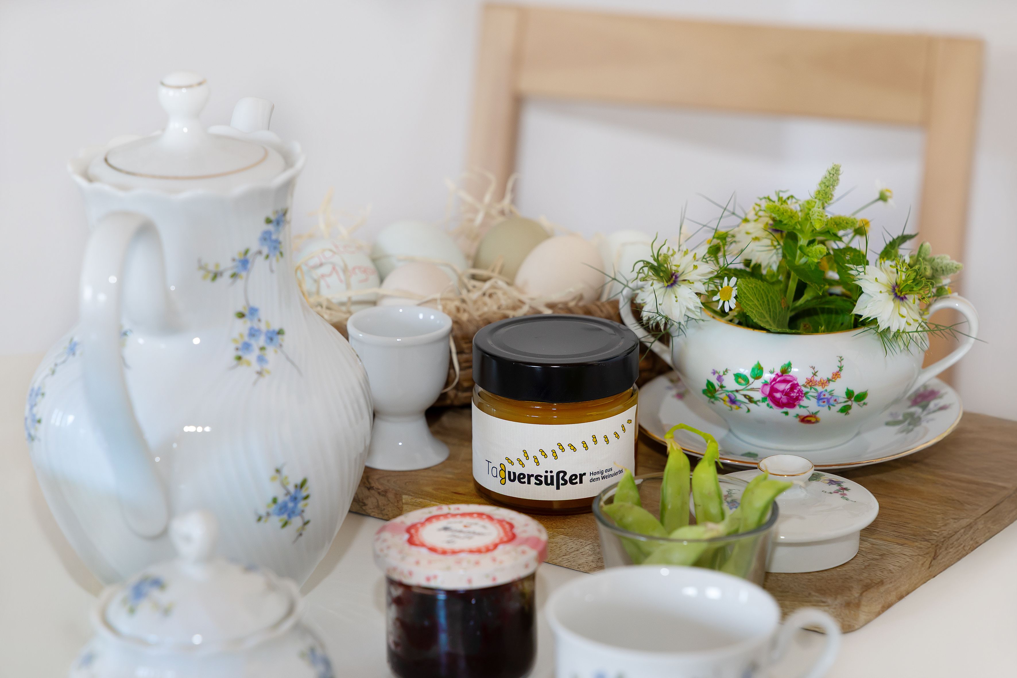 Frühstückstisch mit Teekanne, Honigglas, Marmelade, Eiern und Blumen.