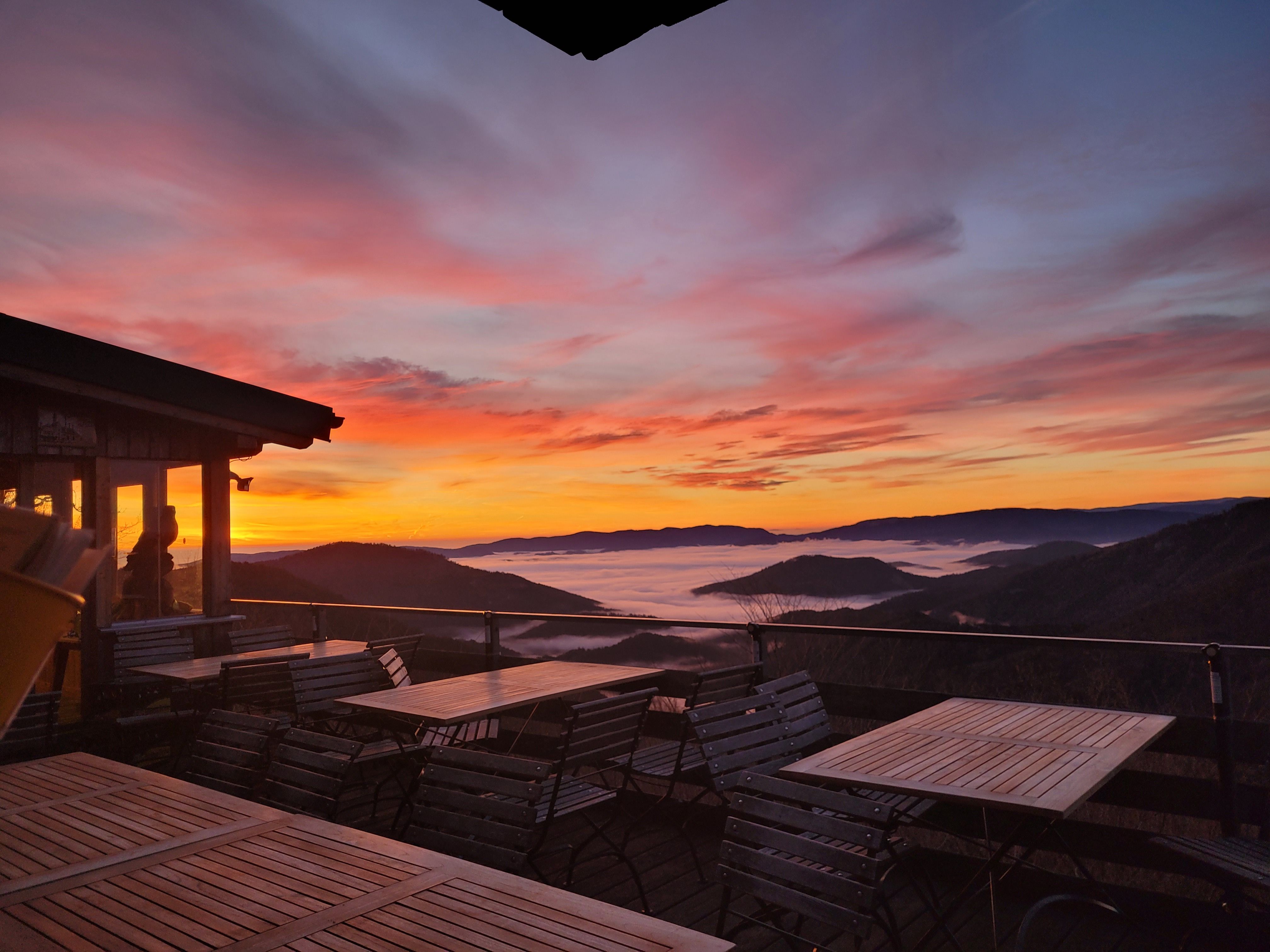 Terrasse der Enzianhütte1 mit Tischen und Stühlen bei Sonnenaufgang über einem Nebelmeer in den Bergen.