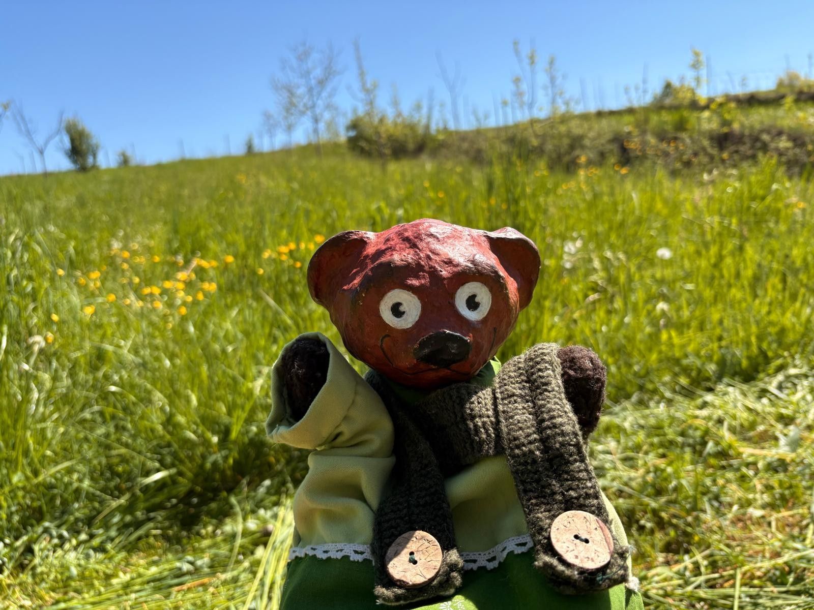 Eine handgefertigte Bärenfigur mit rotem Kopf und großen Augen steht auf einer grünen Wiese unter blauem Himmel.